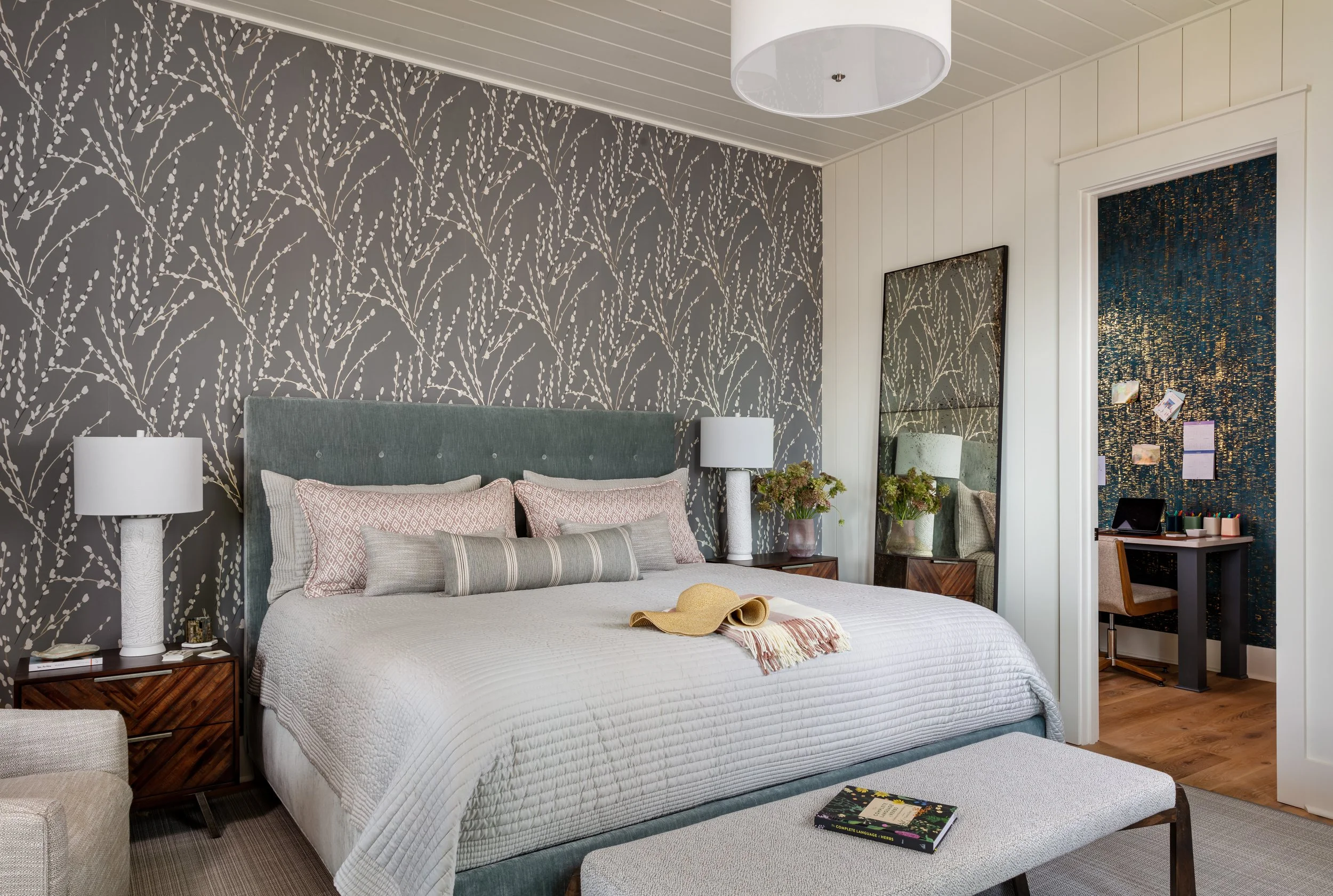 Stylish modern bedroom with patterned wallpaper, upholstered bed, cozy quilt and shams, nightstand lamp & mirrored dresser for chic interior design inspiration.