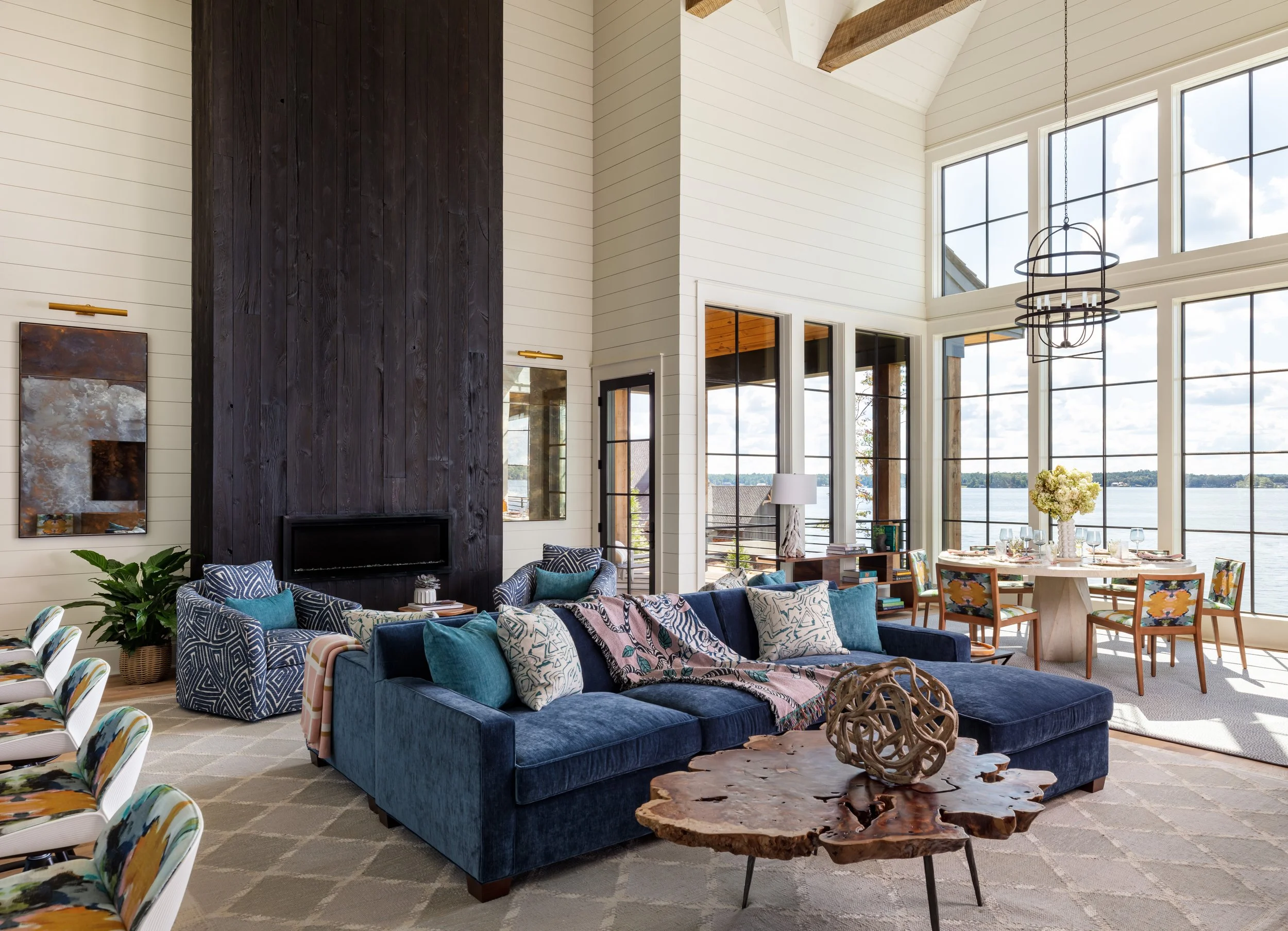 Modern high-ceiling living room with dark wood fireplace accent wall, blue sectional sofa, patterned chairs, large lake-view windows, and rustic coffee table for stylish interior design inspiration