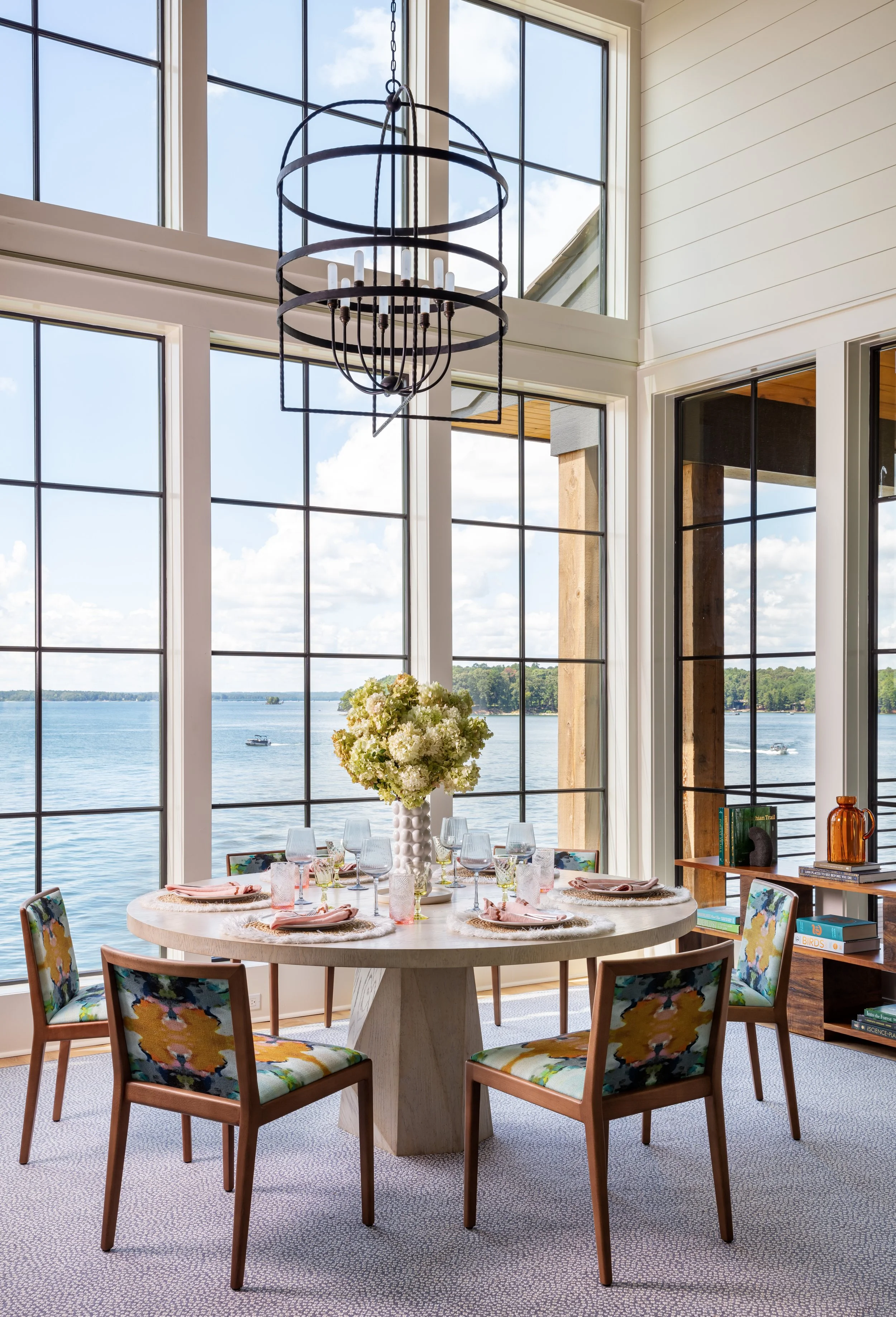 Elegant lakeside dining room with tall windows, modern round table, colorful patterned chairs, floral centerpiece, and sleek black chandelier for stylish interior design inspiration
