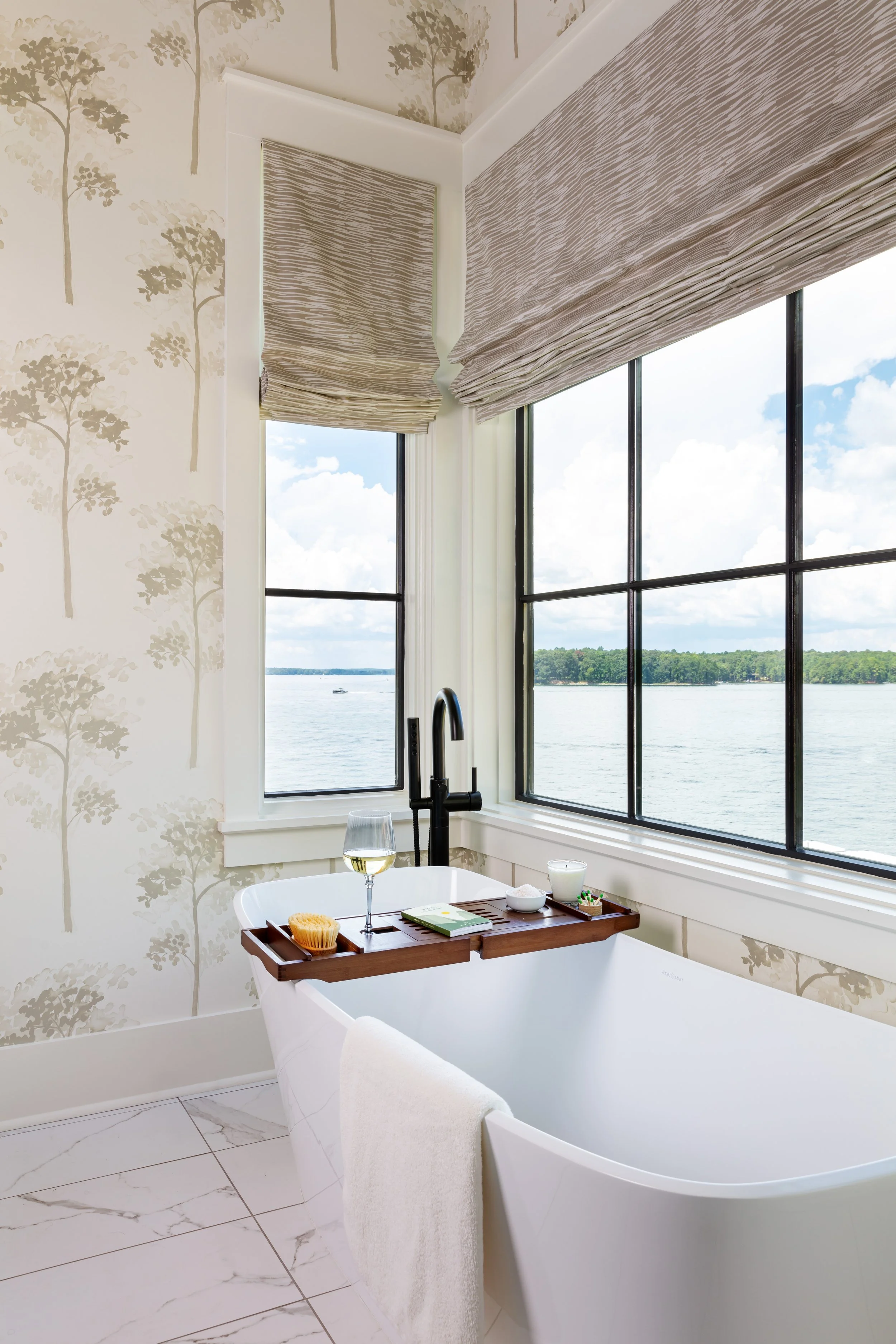 Luxurious freestanding bathtub with wooden bath tray by large windows overlooking water, chic wallpaper & modern faucet for serene spa‑like bathroom design inspiration