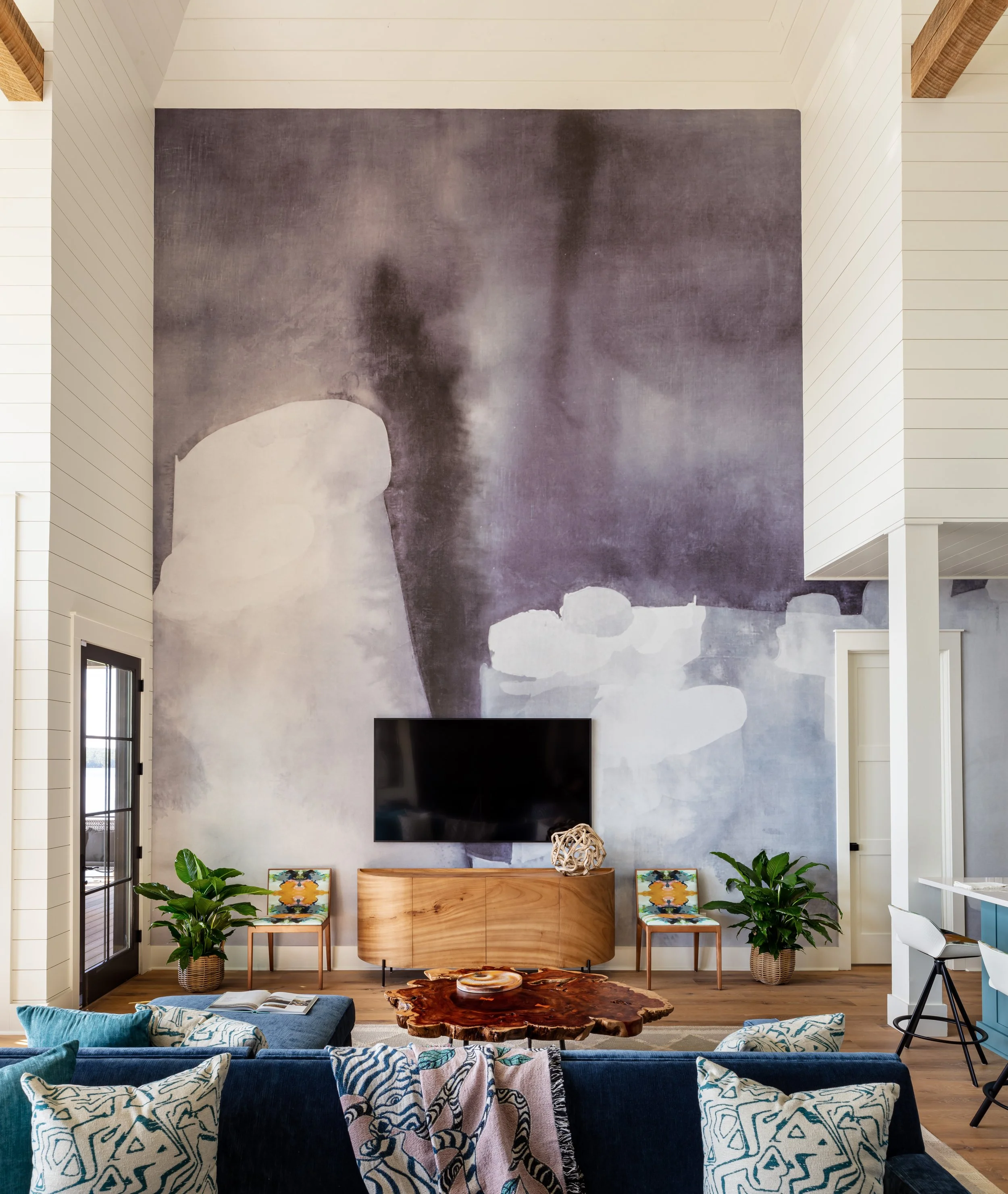 Modern living room with high ceiling, abstract purple-gray wall mural, blue sofa, wooden coffee table, TV on light cabinet, patterned accent chairs & green plants