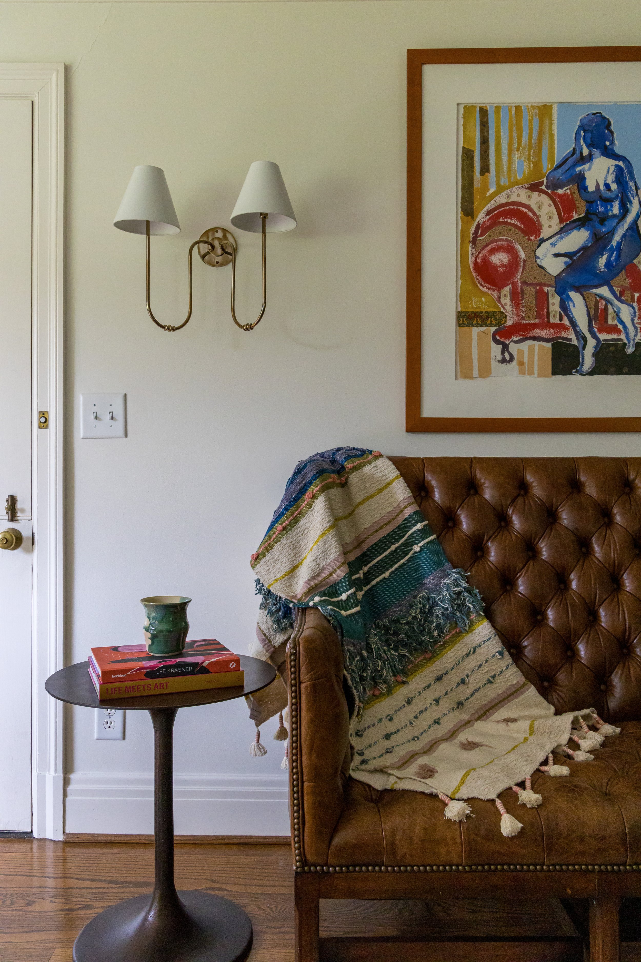 Vintage brown tufted sofa with colorful woven blanket beside a round side table holding books and a green mug, under a brass double sconce and framed abstract art