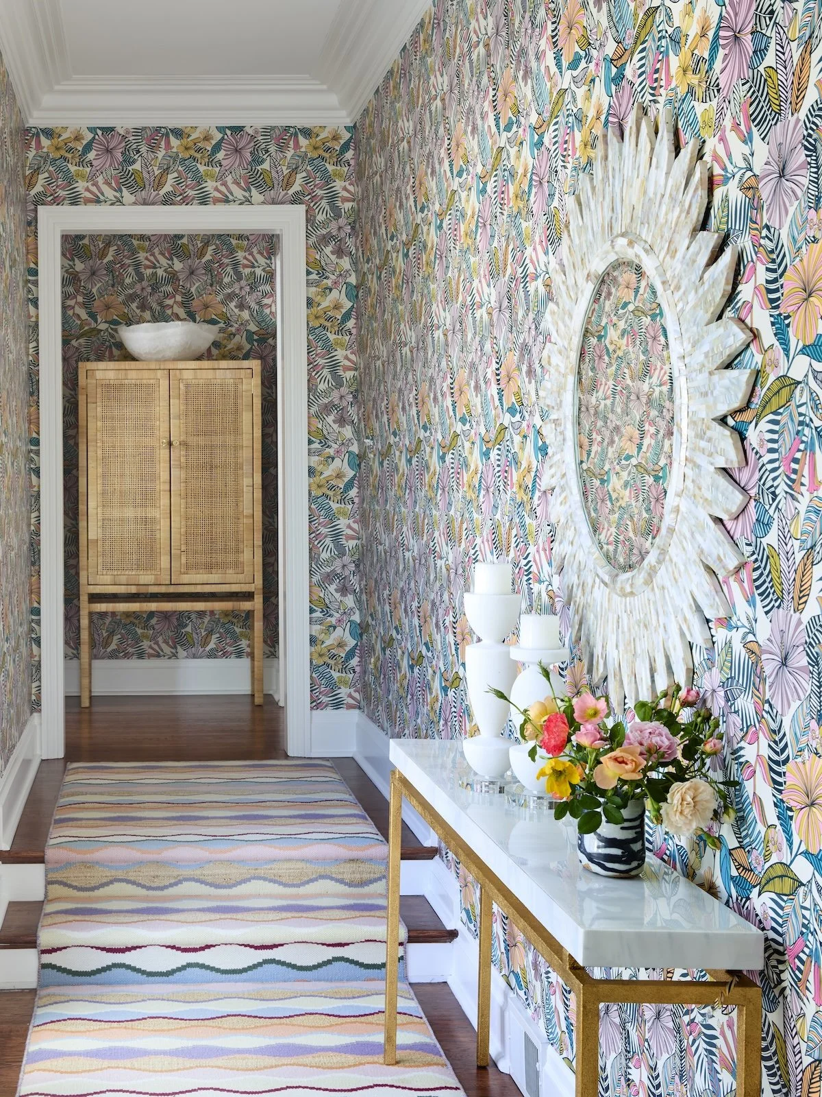 colorful hallway with tropical floral wallpaper mother of pearl sunburst mirror marble console rattan cabinet and multicolor runner by betsy wentz