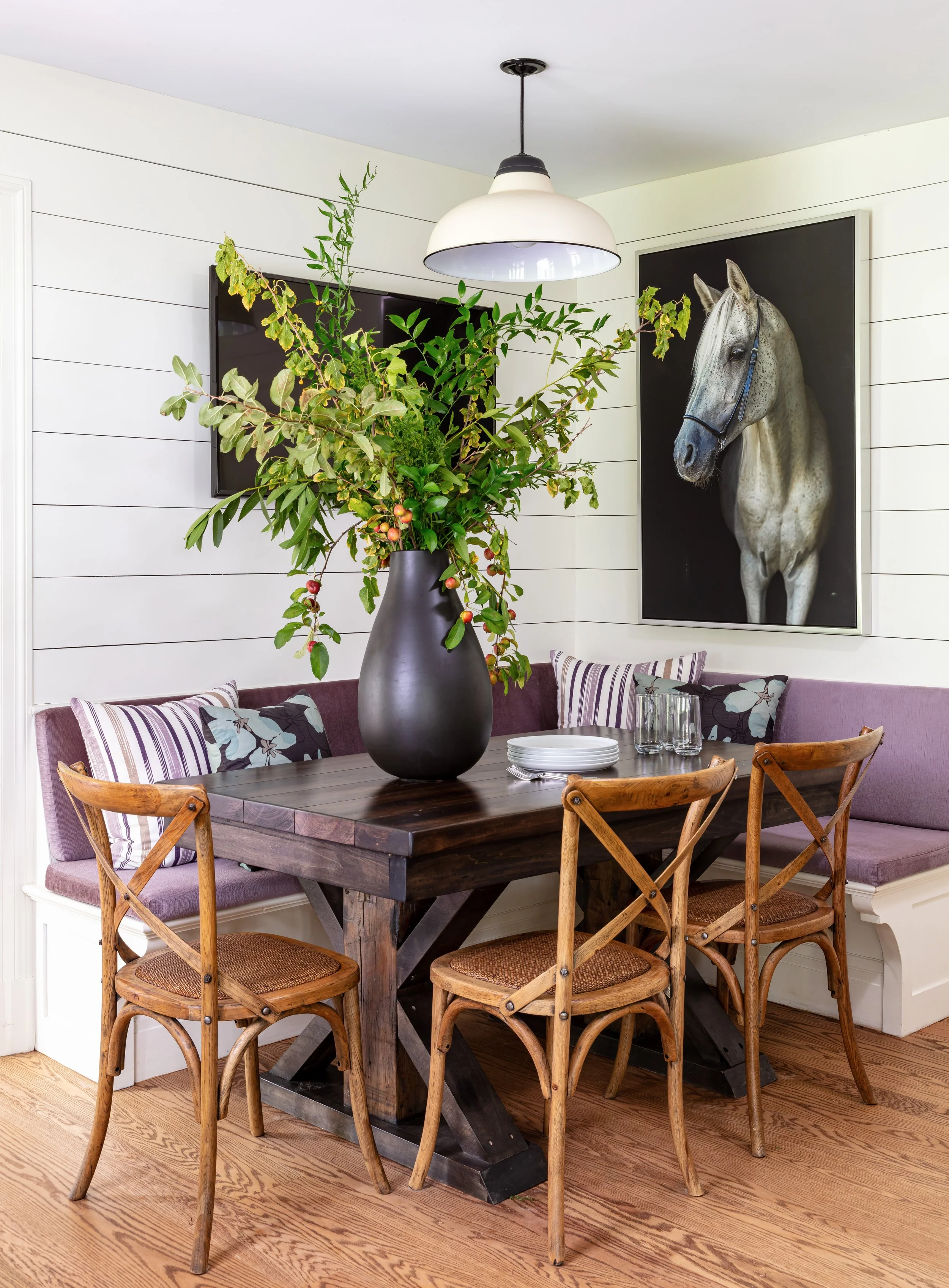 cozy dining nook with shiplap walls, purple banquette, wooden X-back chairs, dark table, greenery vase & horse artwork for cozy farmhouse interior design inspiration