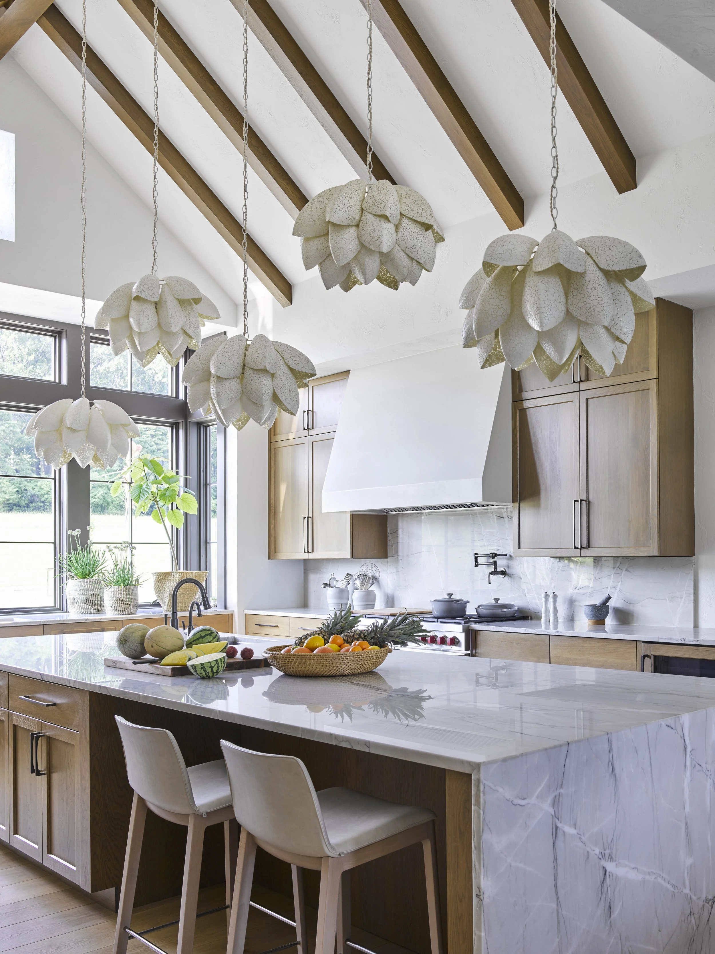 Spacious kitchen with wooden beams, marble island, modern chairs, elegant lights, and fruit bowls. Natural light and greenery add a fresh ambiance.