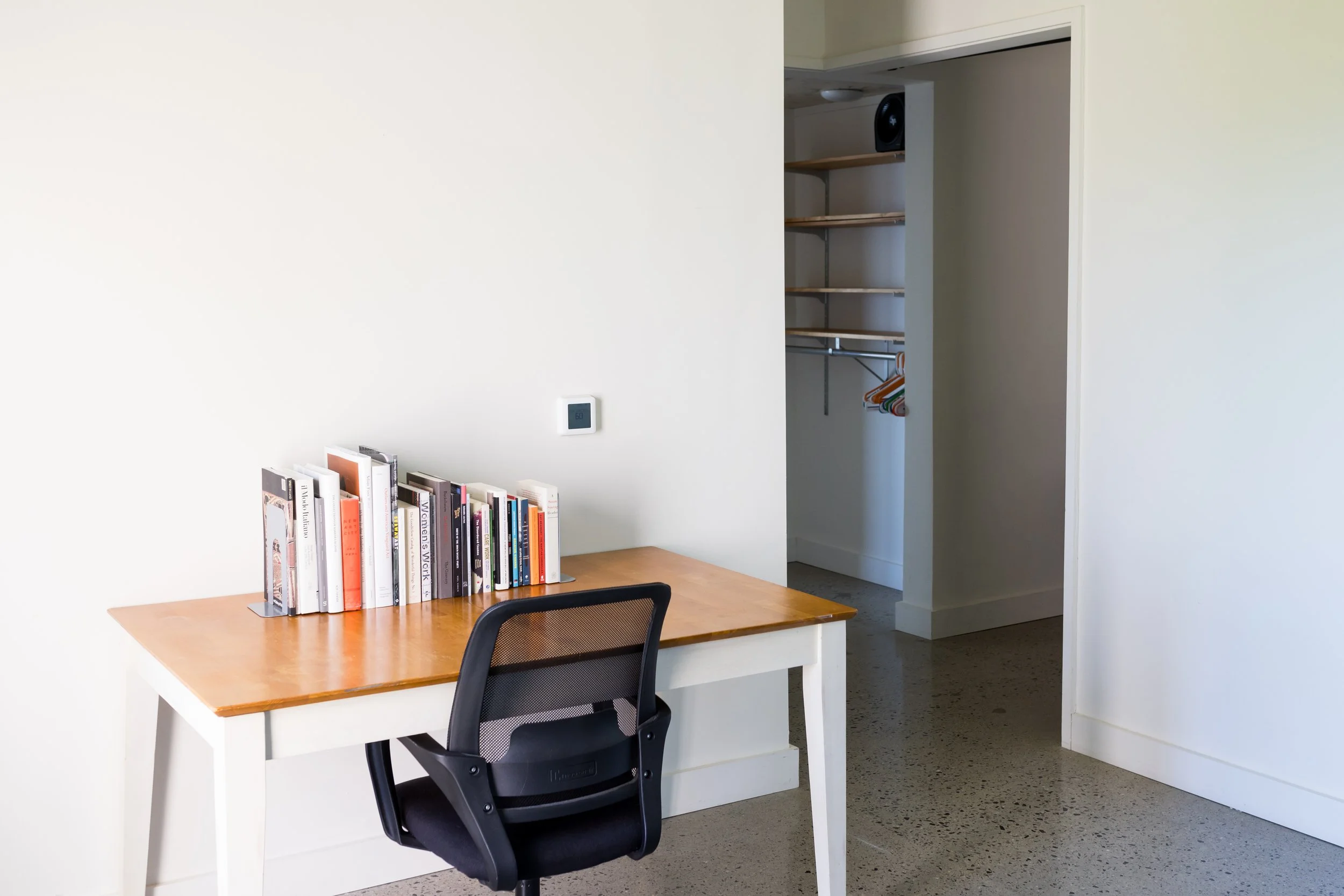 Minimalist home office with wooden desk, books, black office chair, open closet with shelves and hangers.