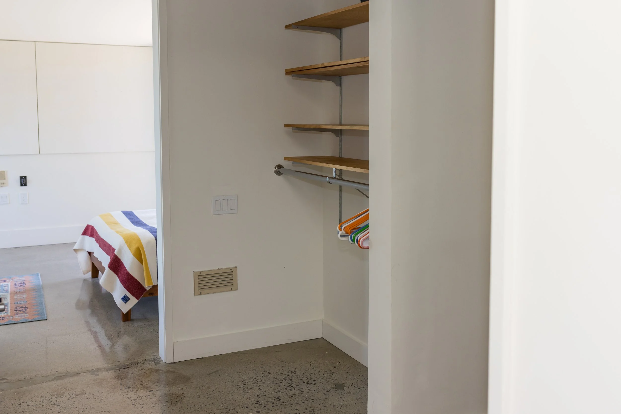 Open closet with wooden shelves and colorful hangers, adjacent to a bedroom with a striped blanket on the bed.