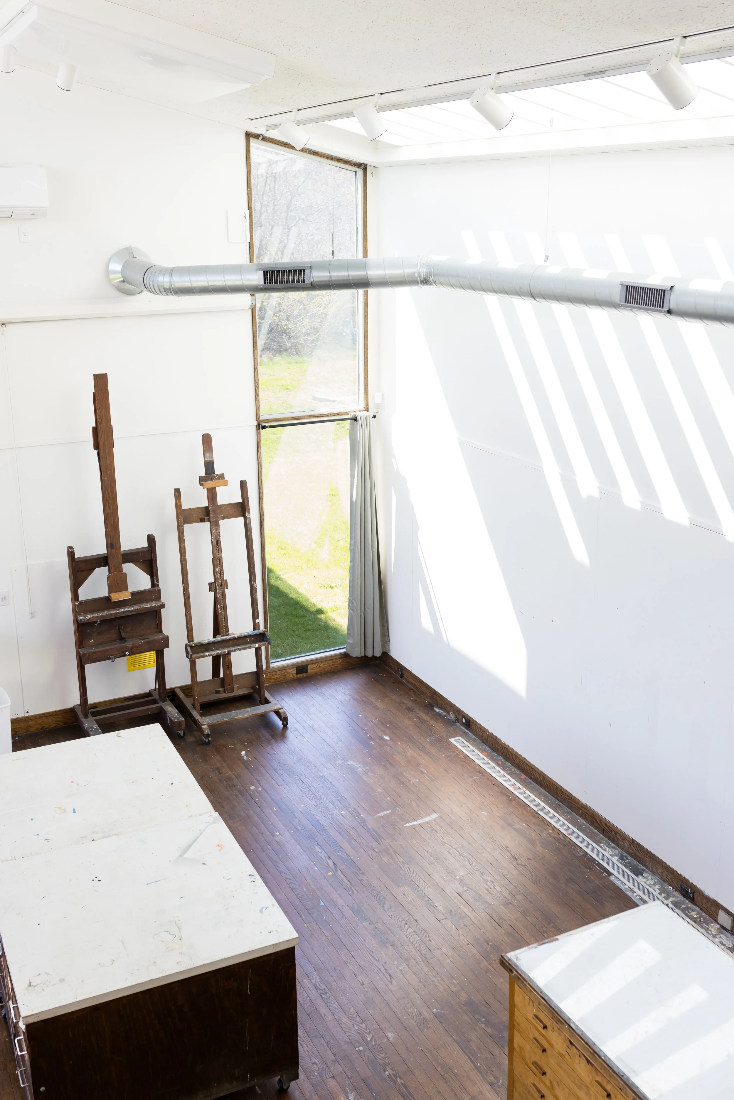 Art studio interior with easels, wooden floor, large window, and overhead ductwork.