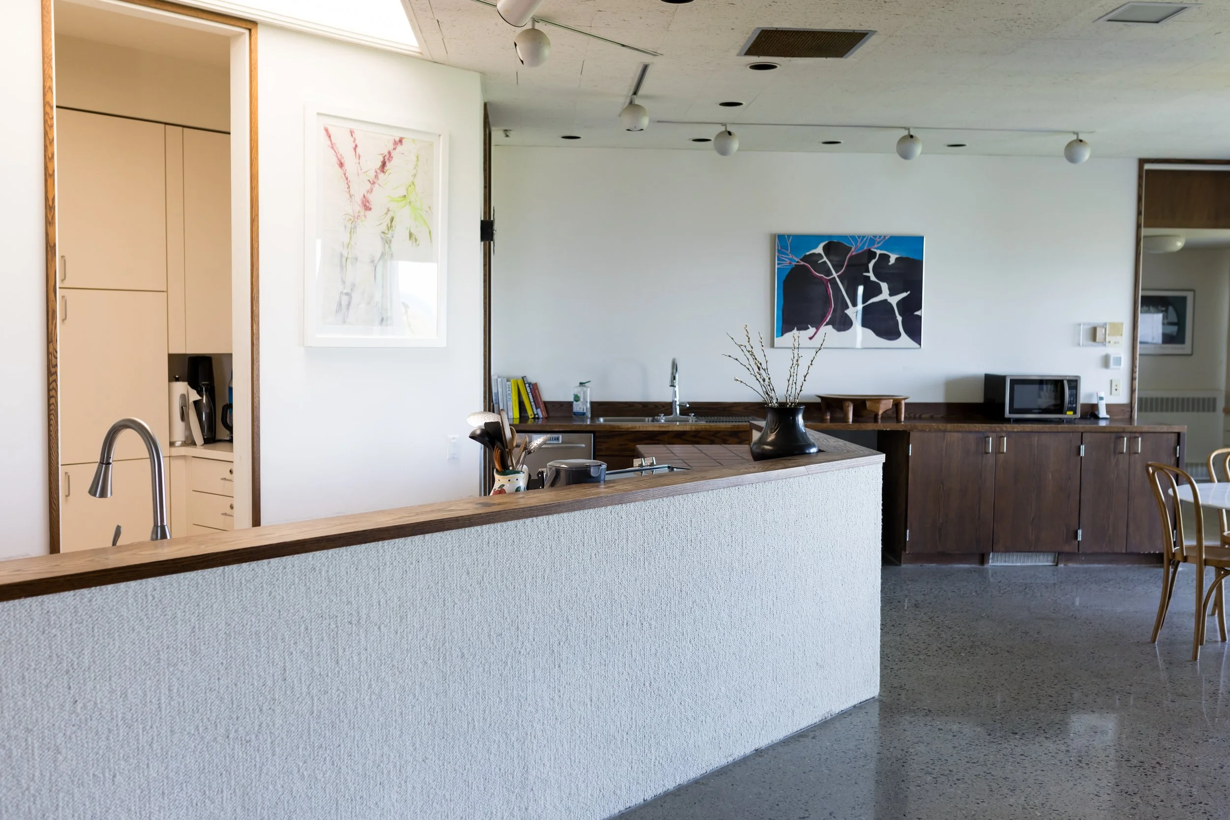 Modern kitchen interior with white walls, a wooden countertop, a sink, a microwave, decorative vase, utensils, and artwork on the walls.