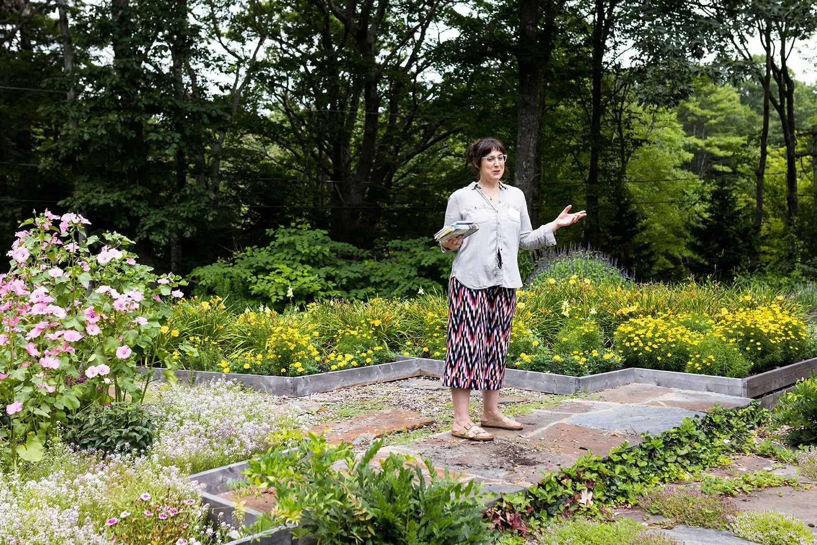 Person standing in a garden with colorful flowers and foliage, wearing patterned skirt and holding notebooks, surrounded by trees.