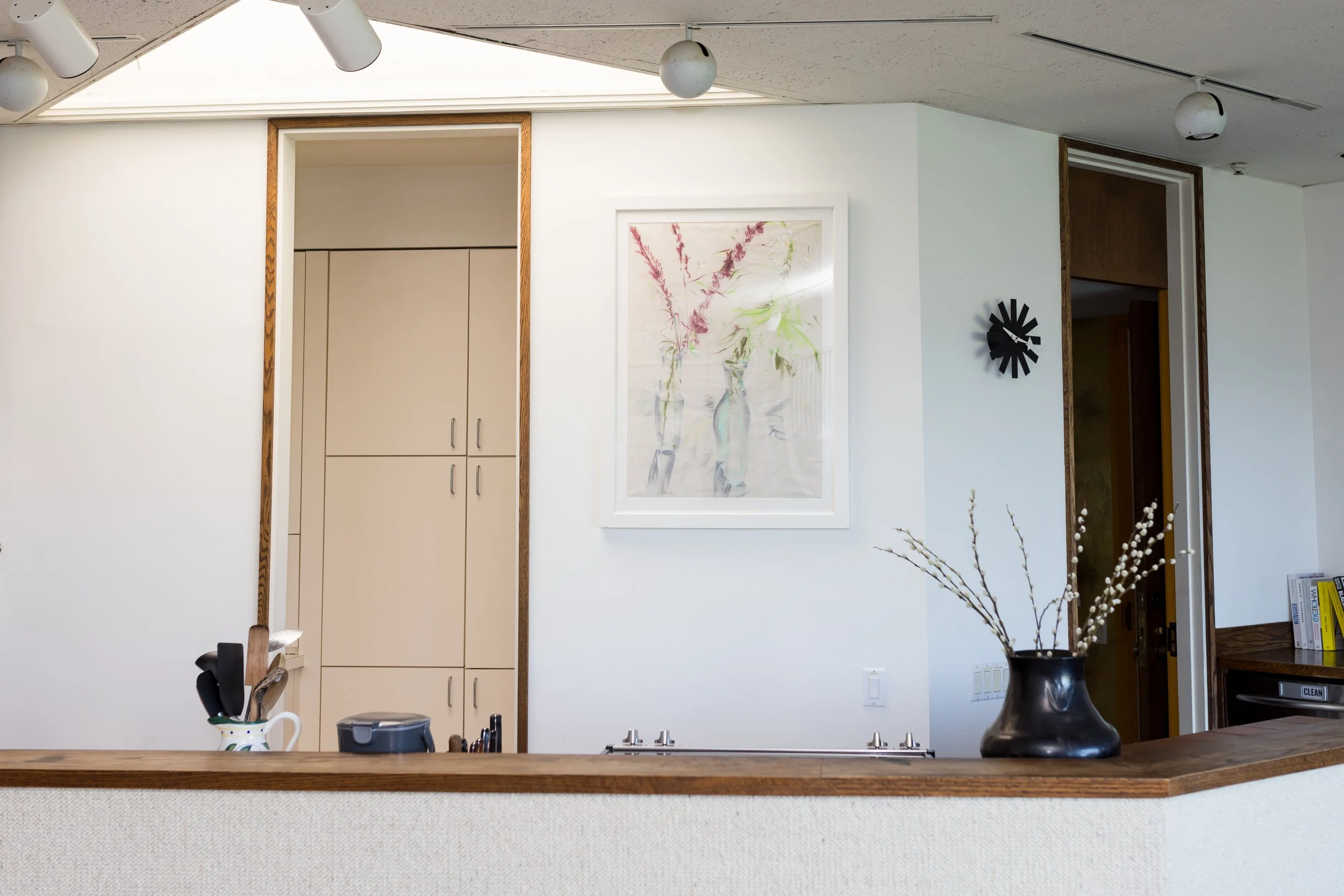 Modern kitchen with light cabinets and utensils in a pitcher, framed artwork on the wall, and a vase with pussy willow branches on a counter.