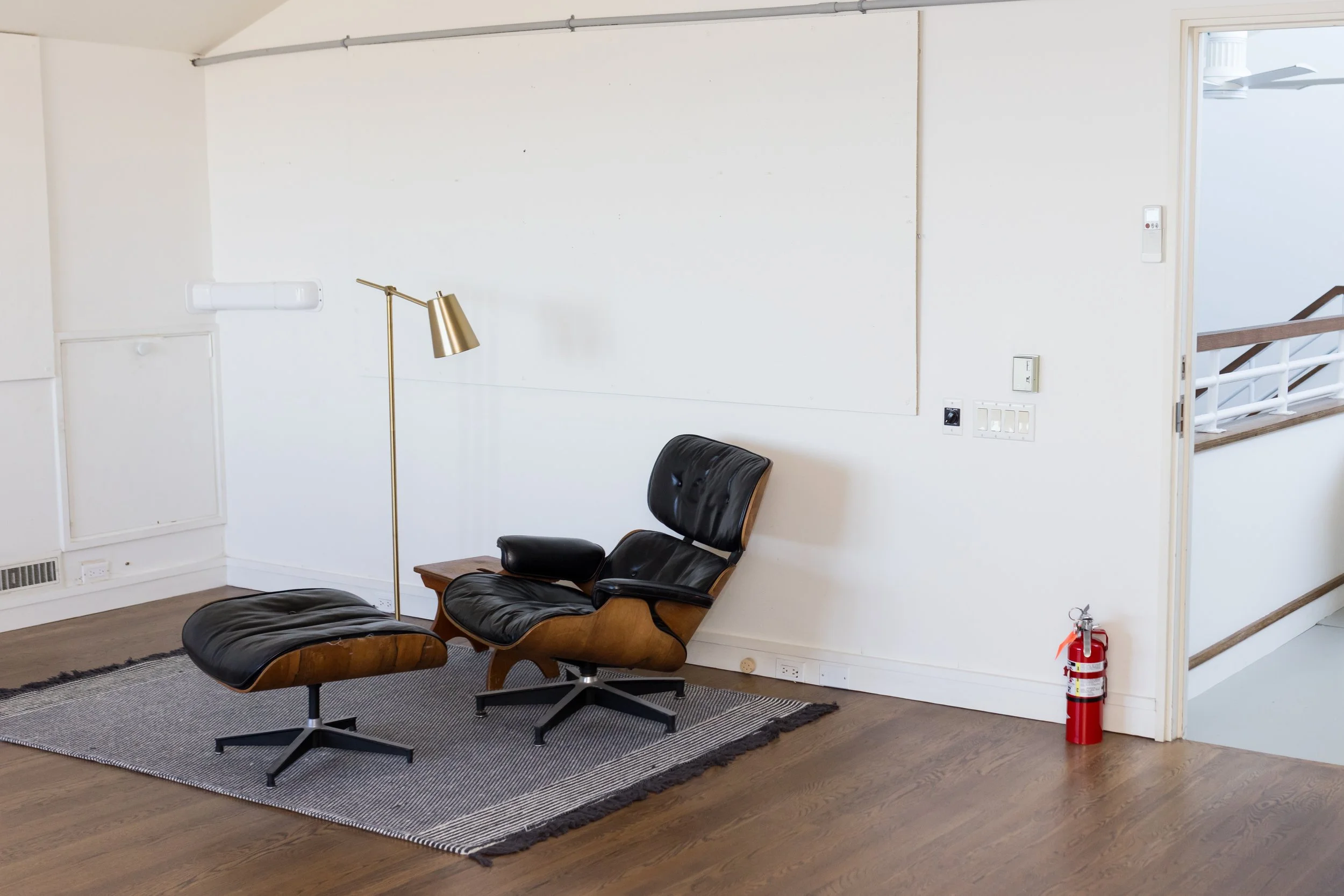 Modern living room with a leather lounge chair and ottoman, floor lamp, small side table, and a fire extinguisher. Wood flooring and a light-colored wall in the background.