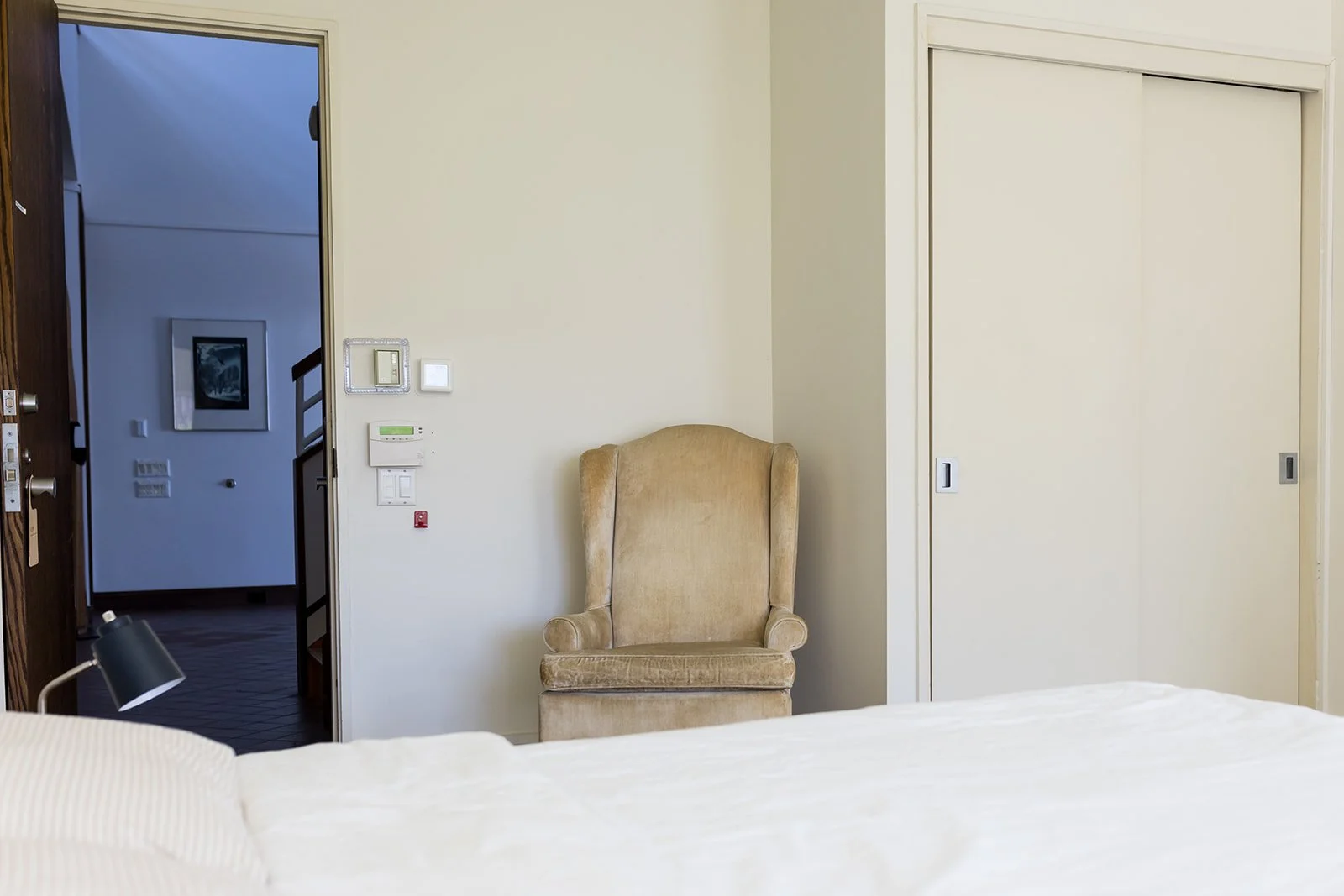 Minimalist bedroom with bed, pillows, bedside lamp, wooden dresser, and a closet and armchair, with the door leading out into the downstairs library common area.