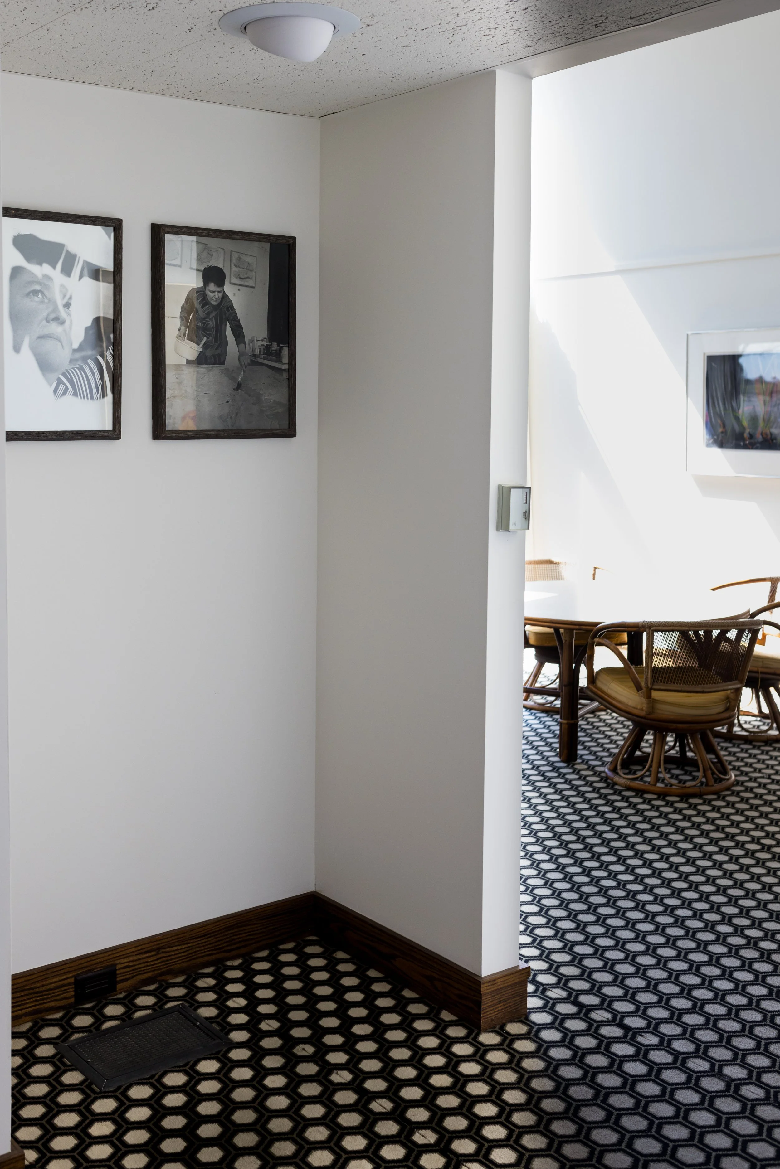 Interior hallway with black and white geometric patterned carpet, framed black and white photographs on the wall, and view into a dining area with rattan chairs and a white table.