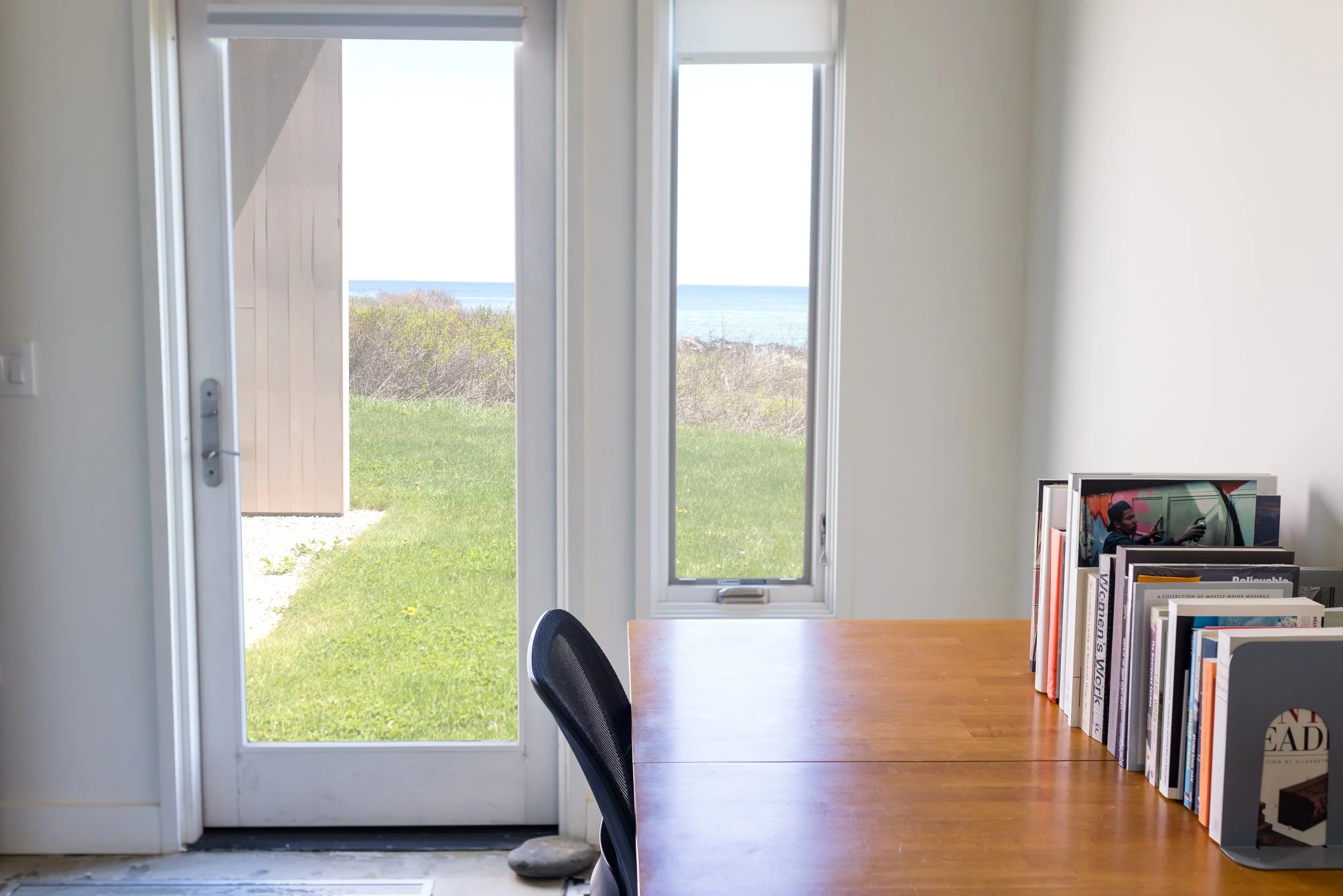 Interior view of a room with a wooden desk and bookshelf, looking out through a glass door to a grassy yard and ocean view.