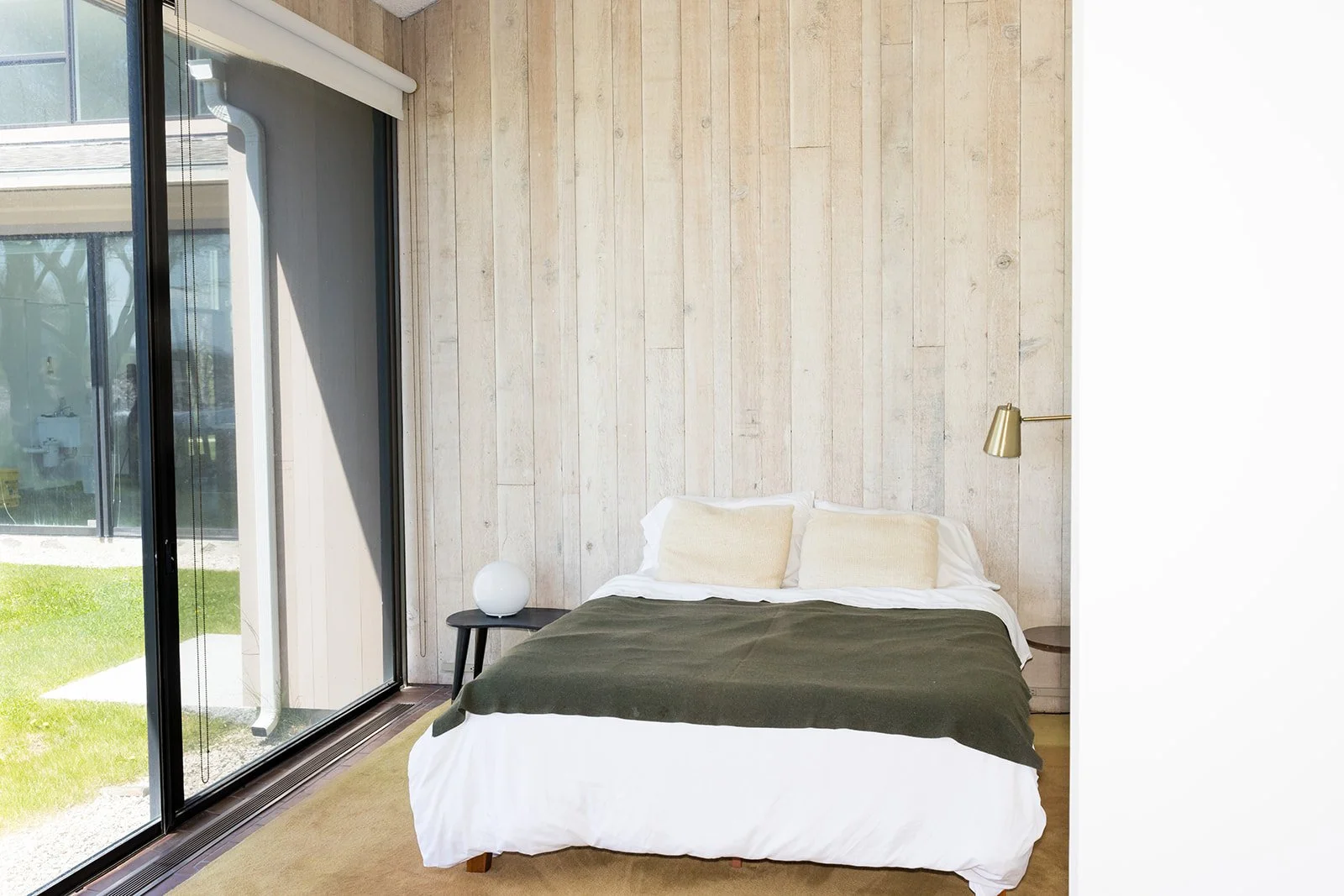Interior of a minimalist room with wooden walls and brown tiled floor, featuring a queen sized bed, a half wall, and large windows with shades.