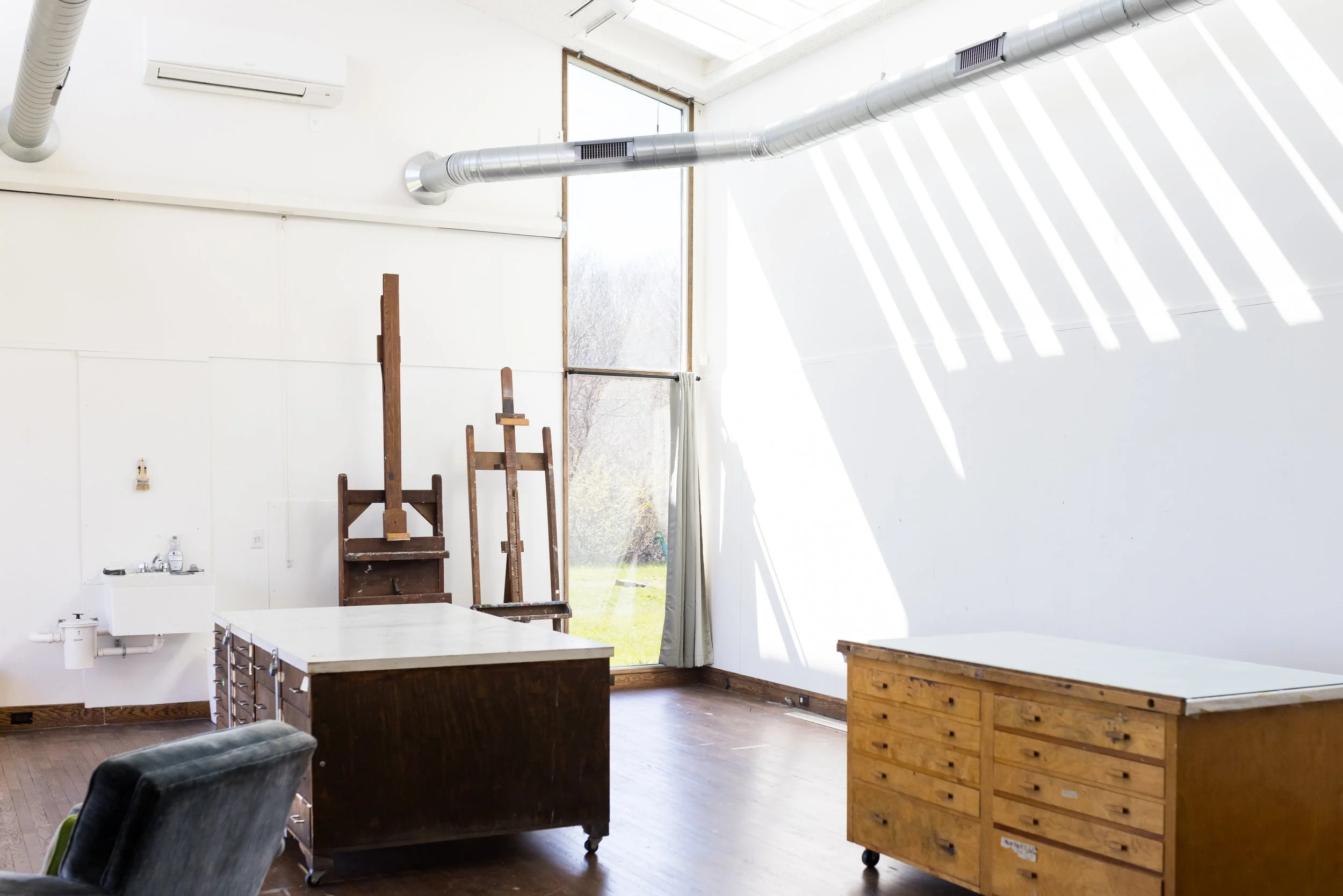 Bright art studio with large windows, wooden easels, work tables, and ventilation ducts.