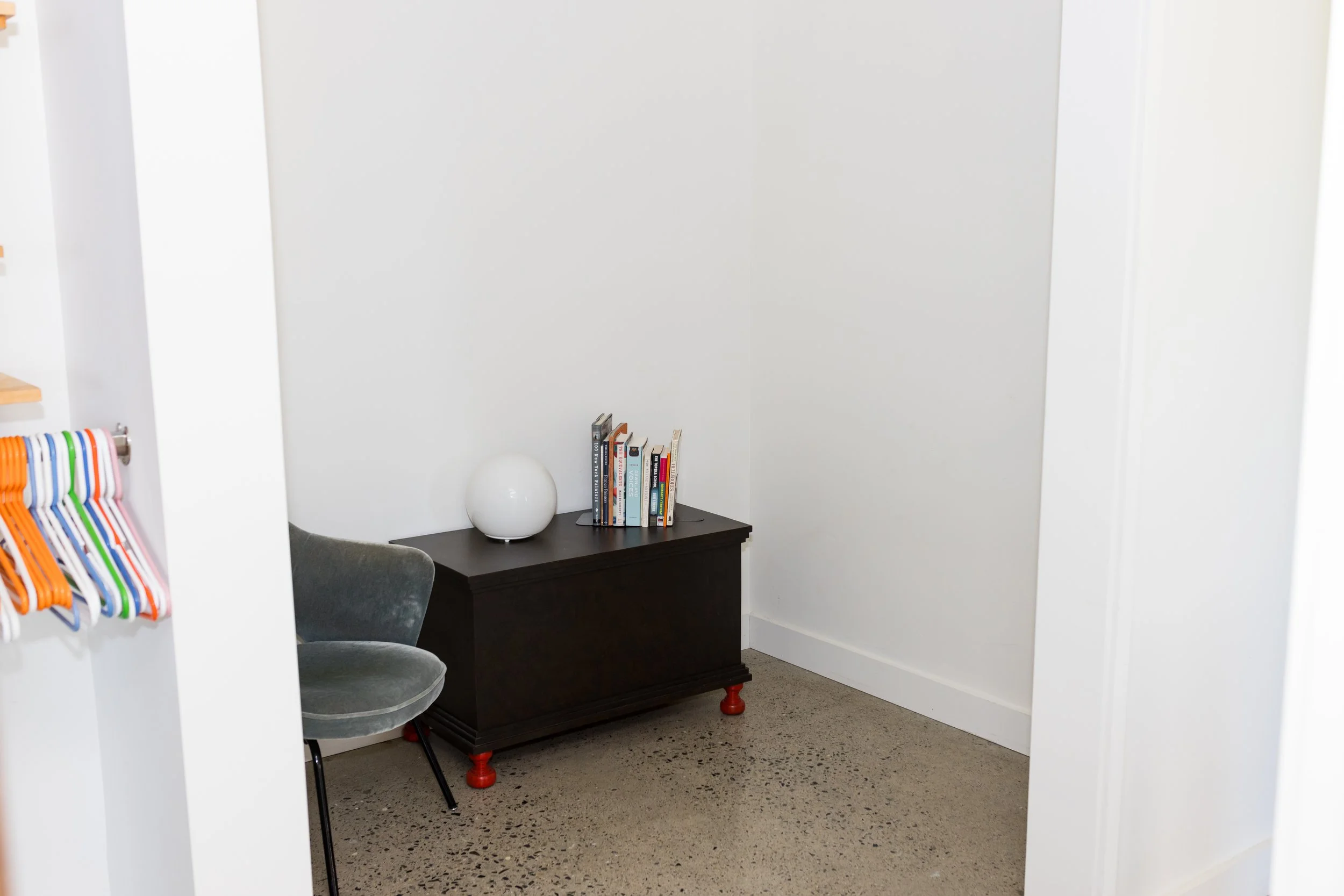 Minimalist room with a gray chair, a wooden cabinet with red feet, a round white lamp, and books. Nearby, colorful hangers are seen on a clothes rack.