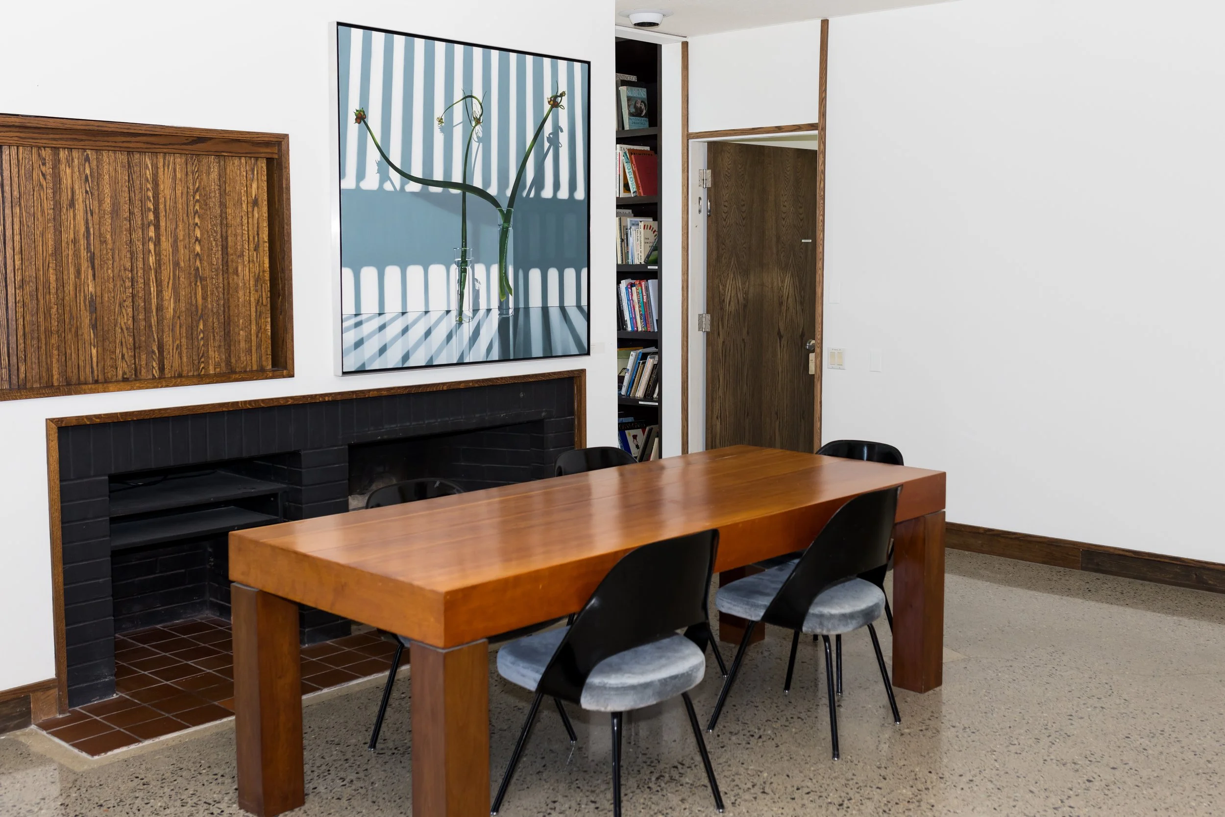 A modern dining room with a wooden table, four black chairs, a fireplace, a framed artwork on the wall, and bookshelves.