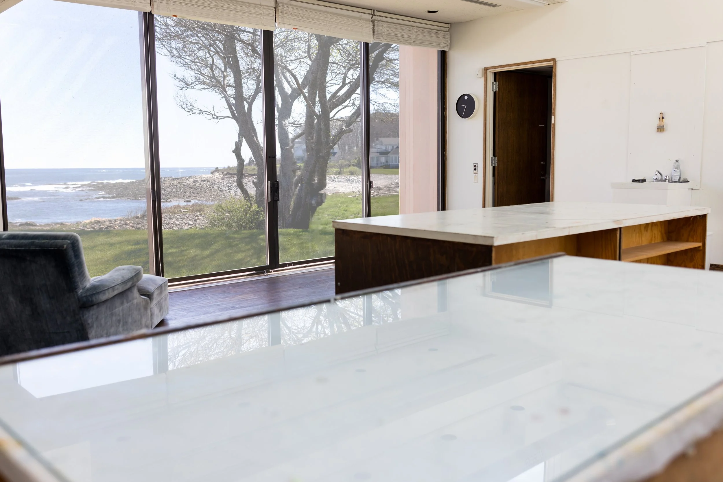 Interior room with large window showing ocean view, trees, and rocky shoreline. Includes a blue armchair, marble-topped tables, and a small sink on the right.