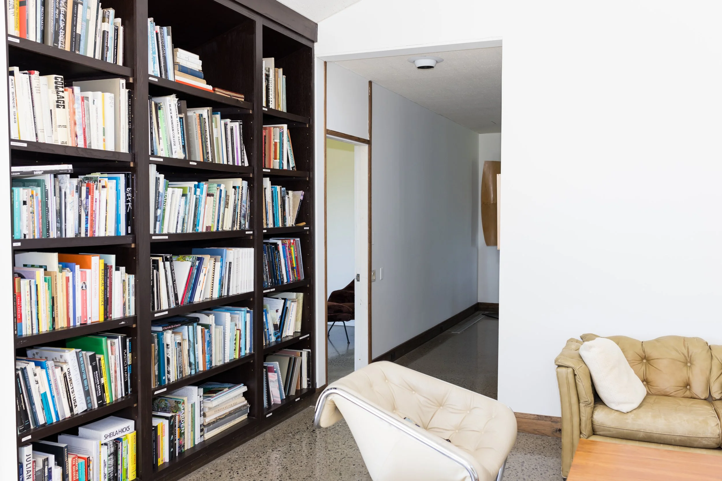 Room with bookshelves filled with books, a beige sofa and armchair, and a hallway in the background.