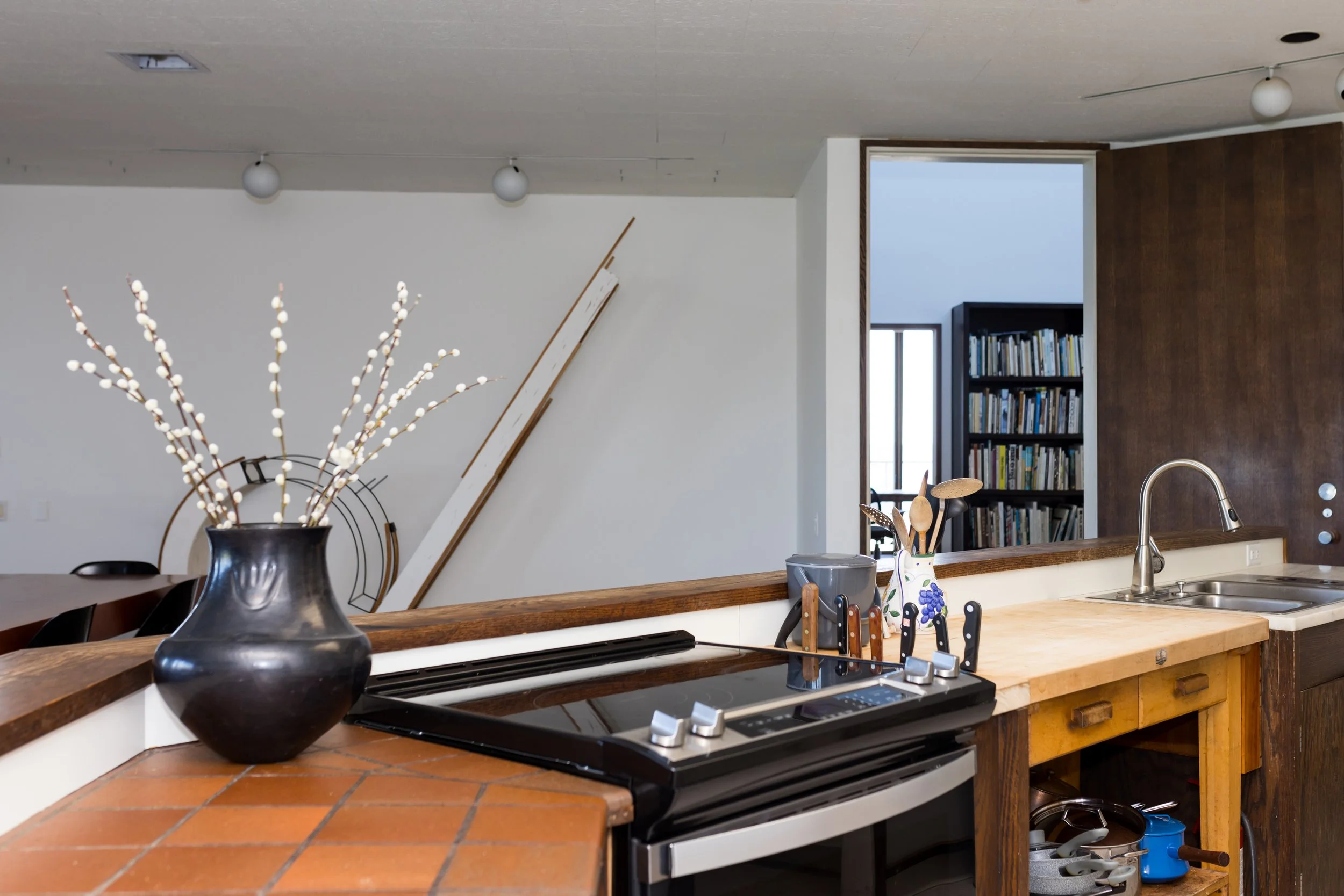 Modern kitchen interior with black vase, electric stove, wooden utensils, and a view of a bookshelf.