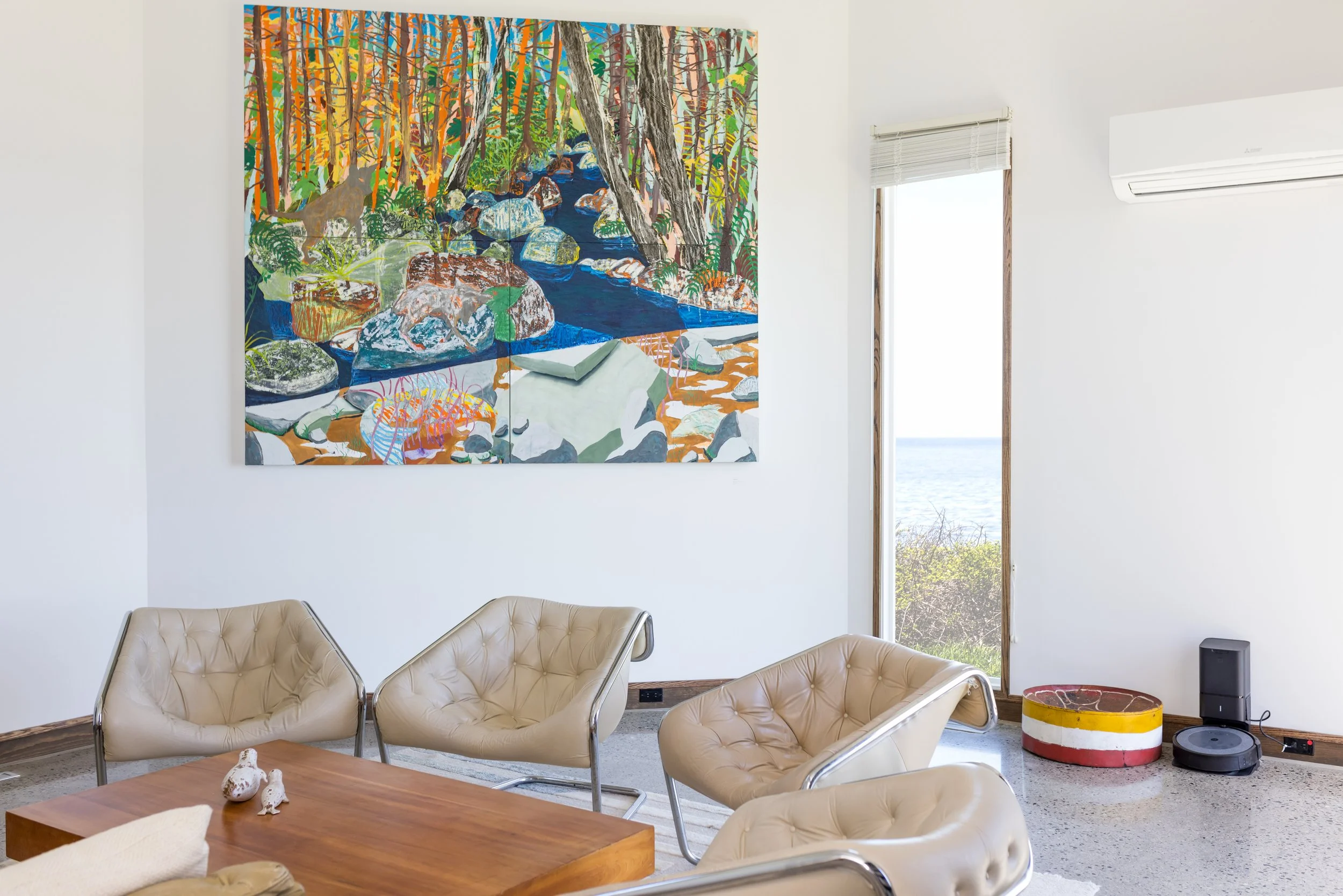 Modern living room with beige chairs, wooden coffee table, large colorful landscape painting on the wall, and a view of a lake through the window.