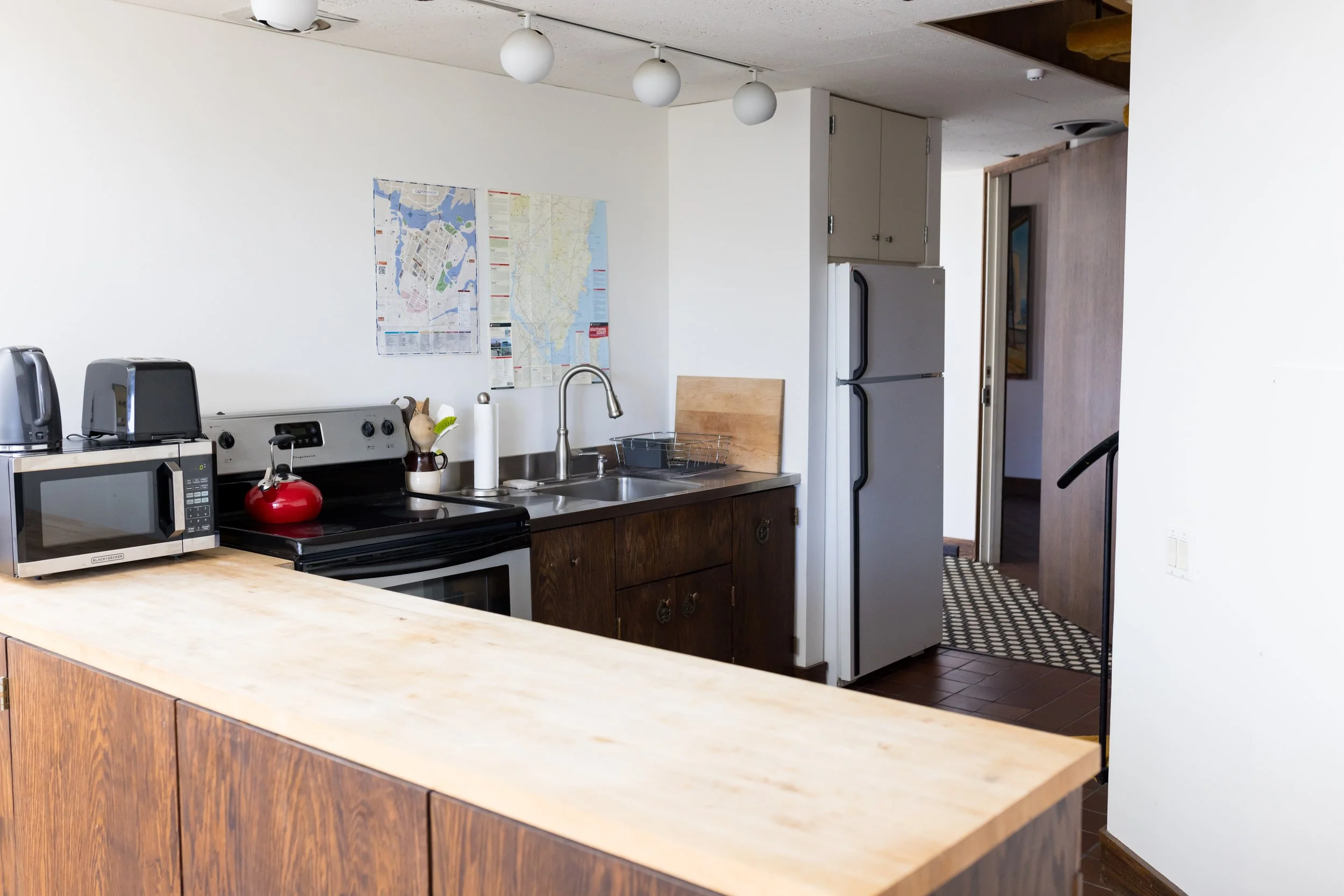 Modern kitchen with wooden cabinets, a microwave, toaster, and kettle on the counter, a stainless steel sink, refrigerator, oven, and wall maps.