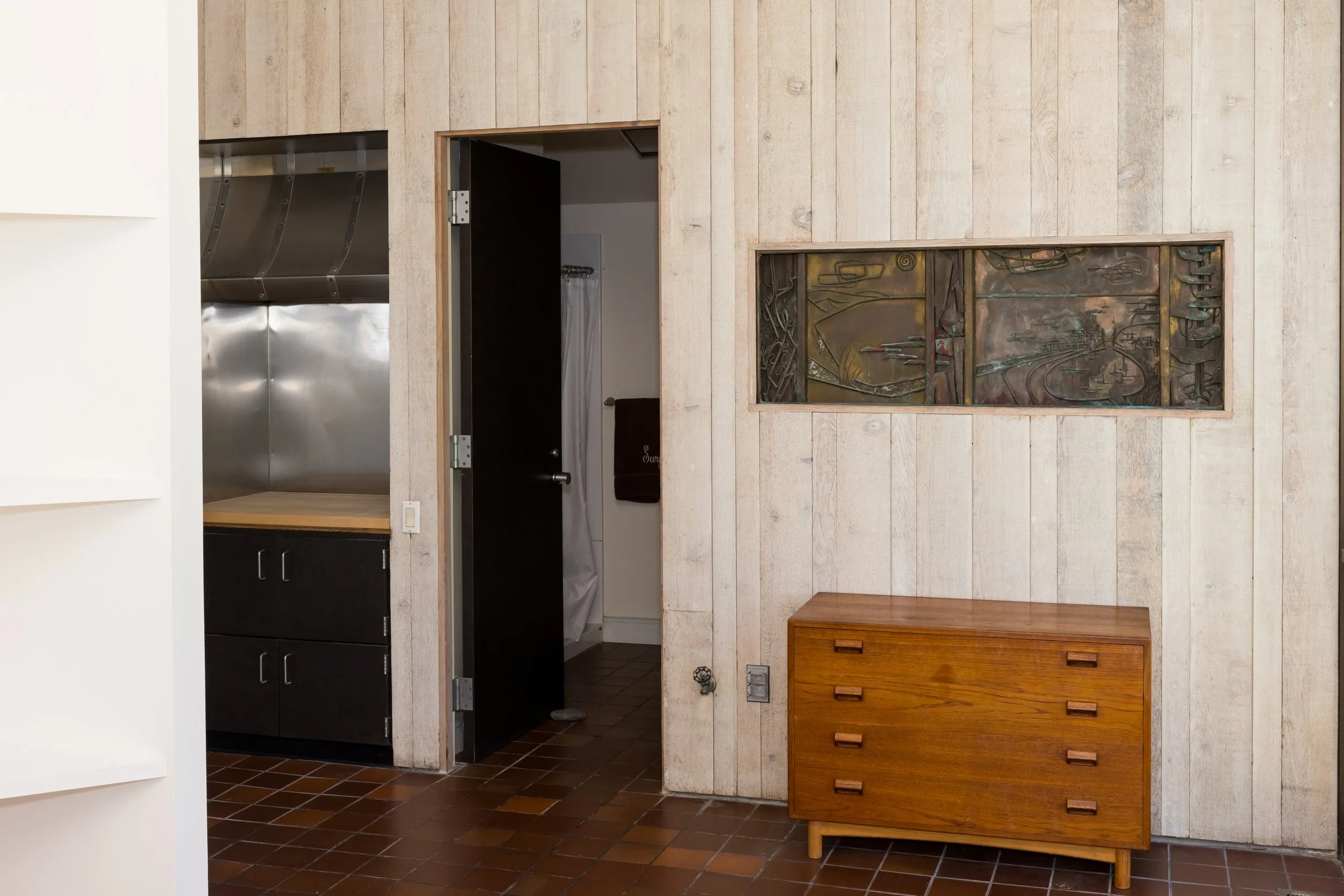 Interior view of a room with a wooden wall, partial open door, metal kitchen counter, tiled floor, and a wooden chest of drawers.