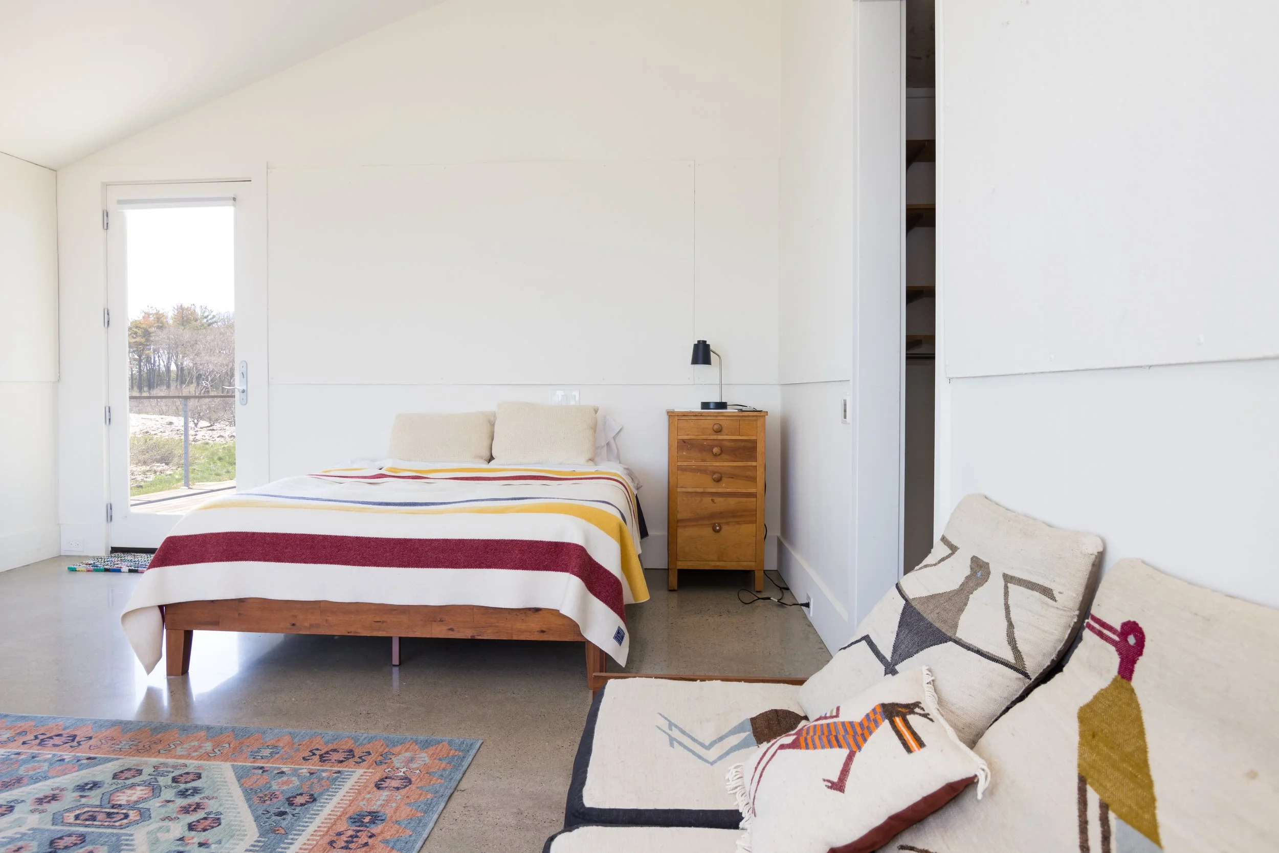 Minimalist bedroom with white walls, a bed with striped blanket, a wood nightstand, and a light fixture. There is a couch with decorative pillows and a patterned rug in the room.