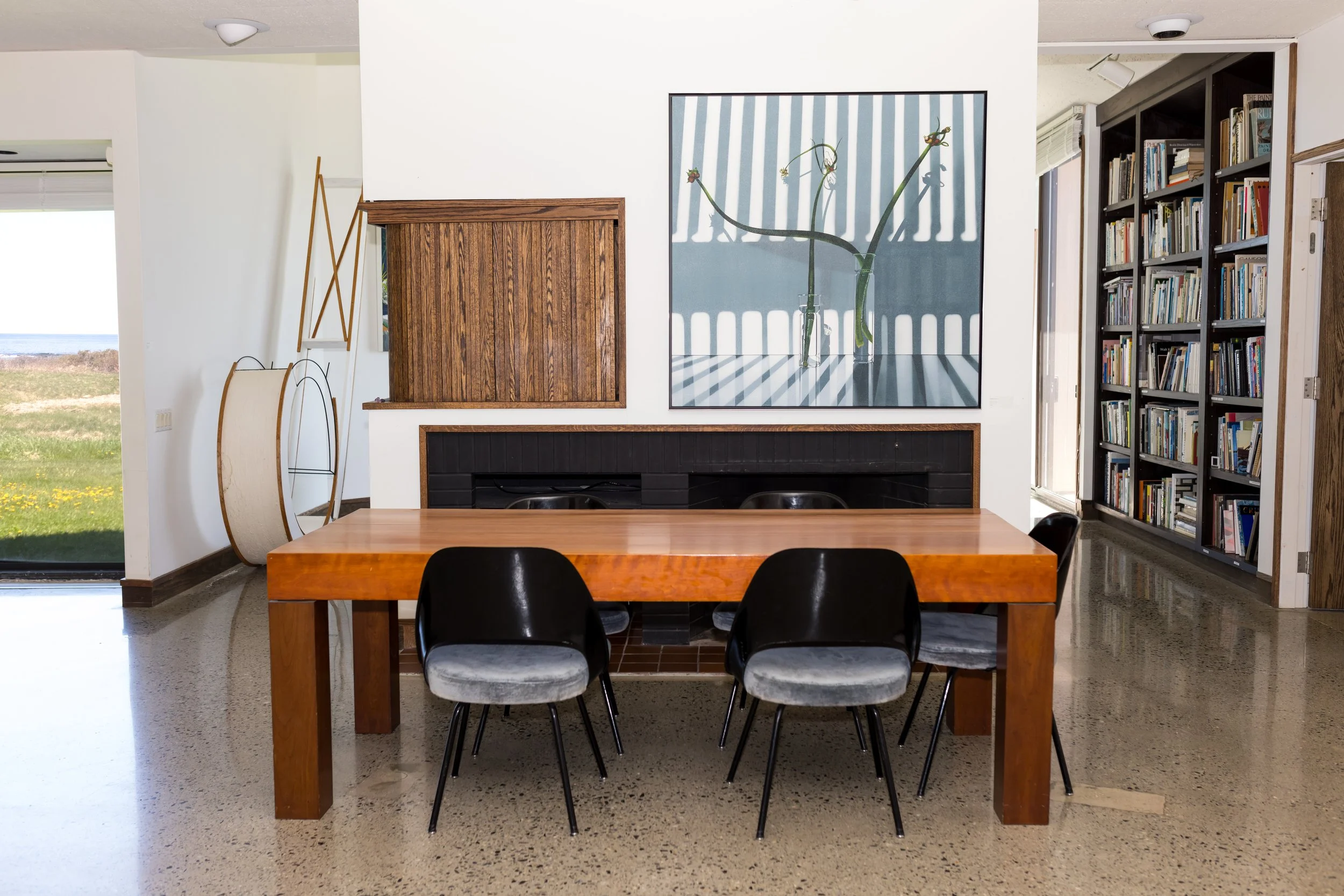 Modern dining room with wooden table, black chairs, and framed artwork on wall, bookshelf in corner, and outdoor view.