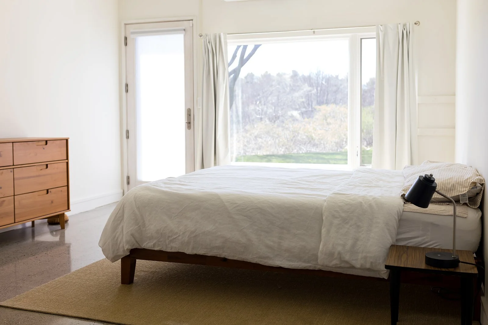 Minimalist bedroom with bed, pillows, bedside lamp, wooden dresser, door leading to the outdoor lawn, and a large window.