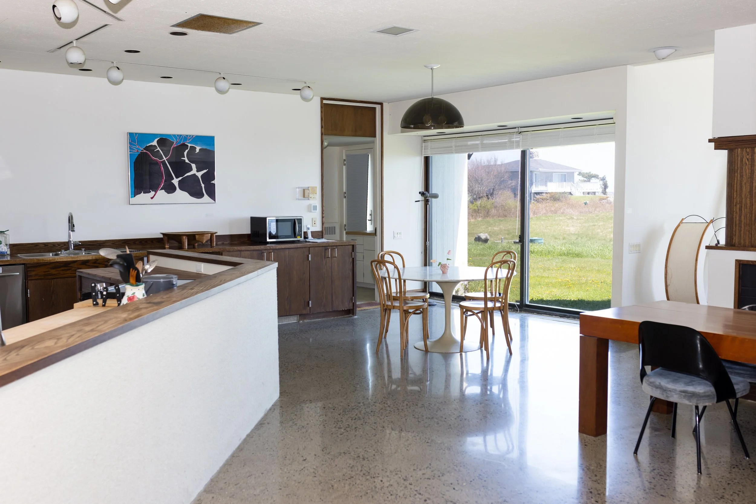 Modern kitchen with round table, wooden chairs, large window, and patio view. Features a countertop with utensils, microwave, and artwork on the wall.