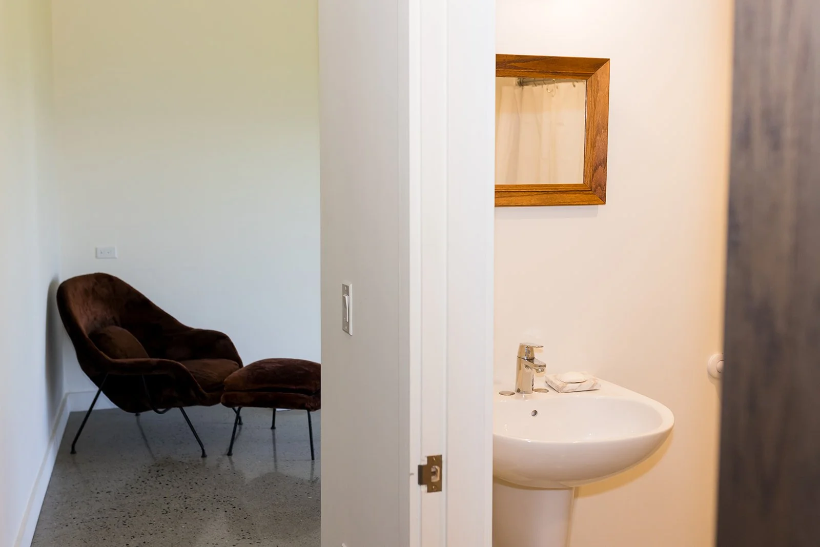 View in hallway of minimalist studio, with bathroom on the right, looking into a sitting area with a comfortable chair and ottoman.