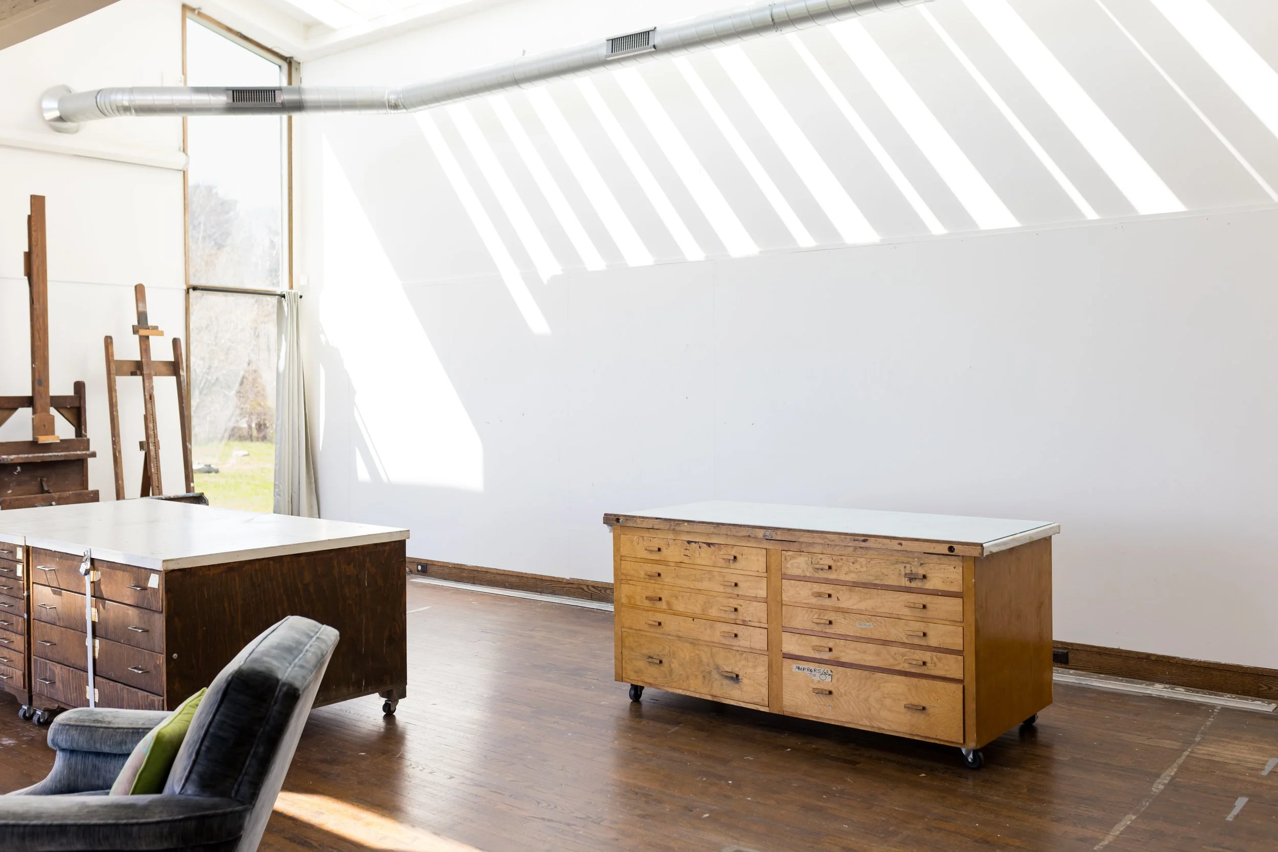 Art studio with rolling wooden cabinets, wooden easels, and a chair on a wooden floor, featuring large windows and angled light from a skylight.