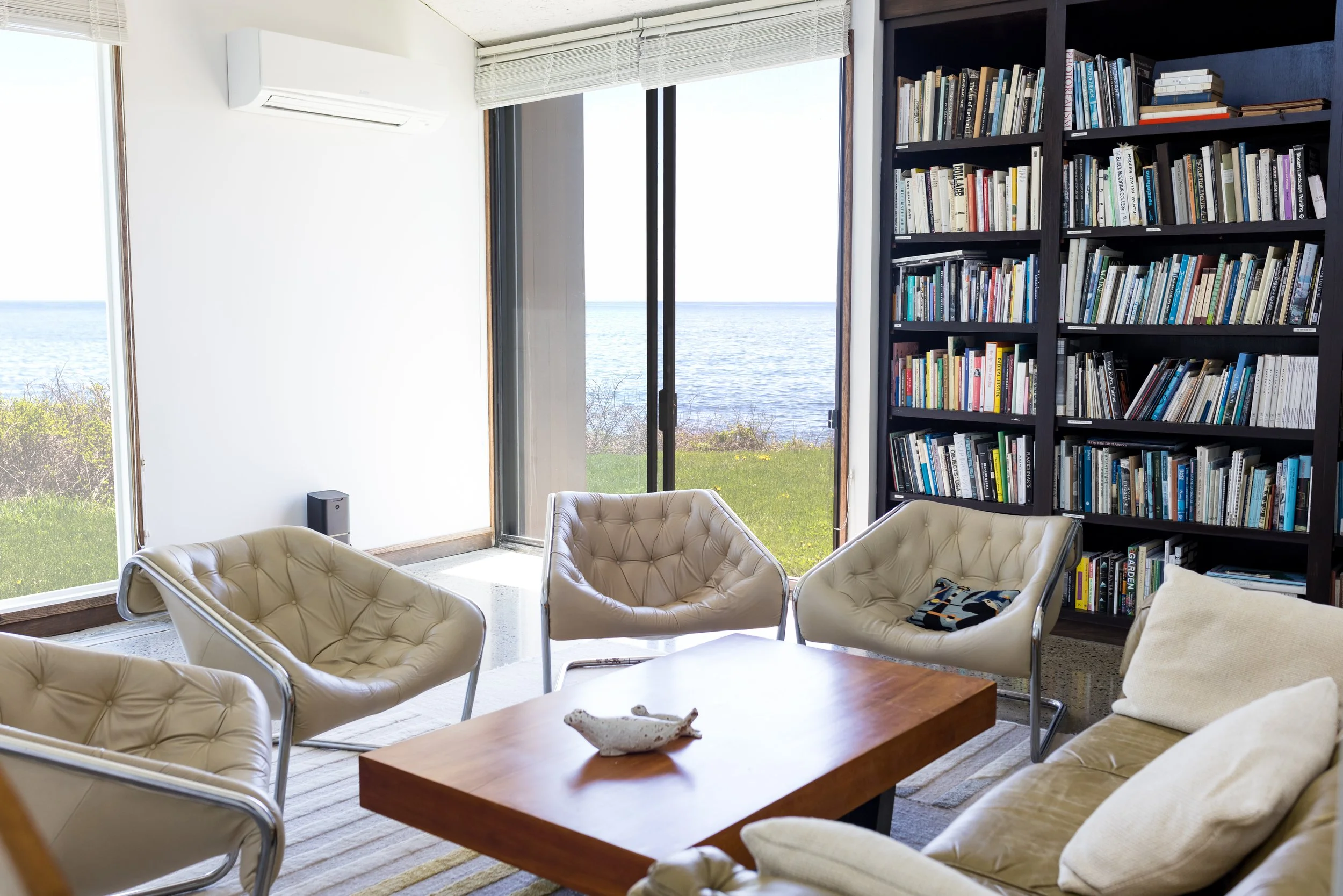 Room with modern seating, bookshelves, and ocean view through large windows.