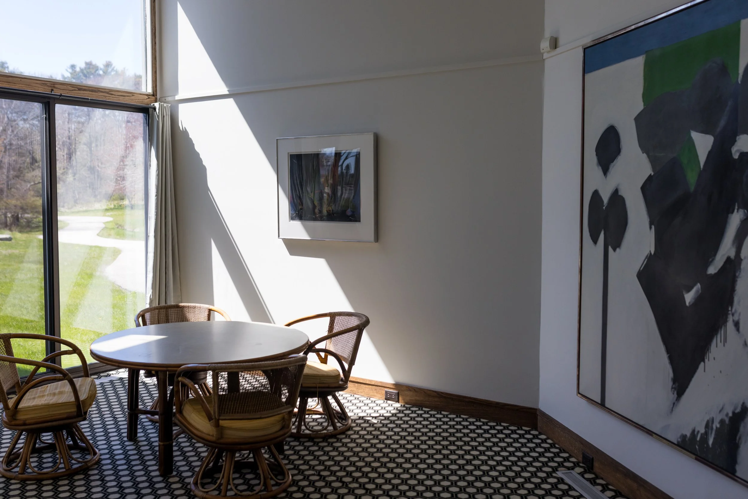 Modern dining area with round table, wicker chairs, abstract wall art, and large window with a garden view.