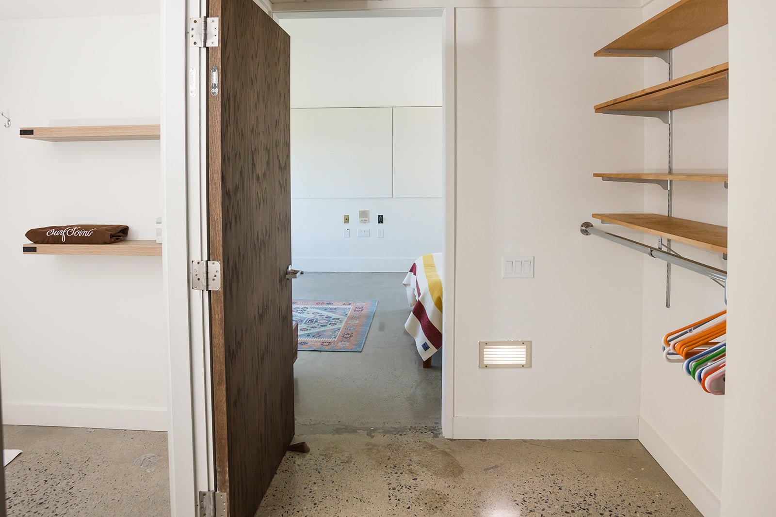 View in hallway of minimalist studio, with bathroom on the left and an alcove with wall mounted shelving on the right, looking into a bedroom.