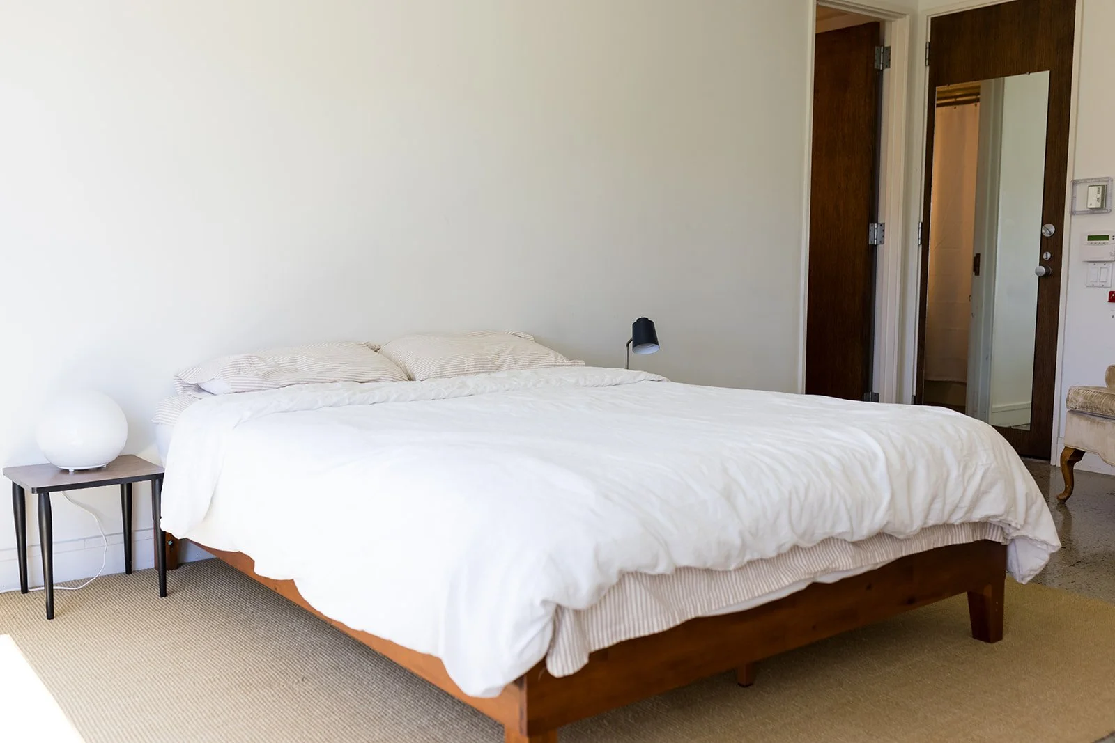 Minimalist bedroom with bed, pillows, bedside lamp, and two doors, one leading to an attached bathroom and one leading to the downstairs library common area.