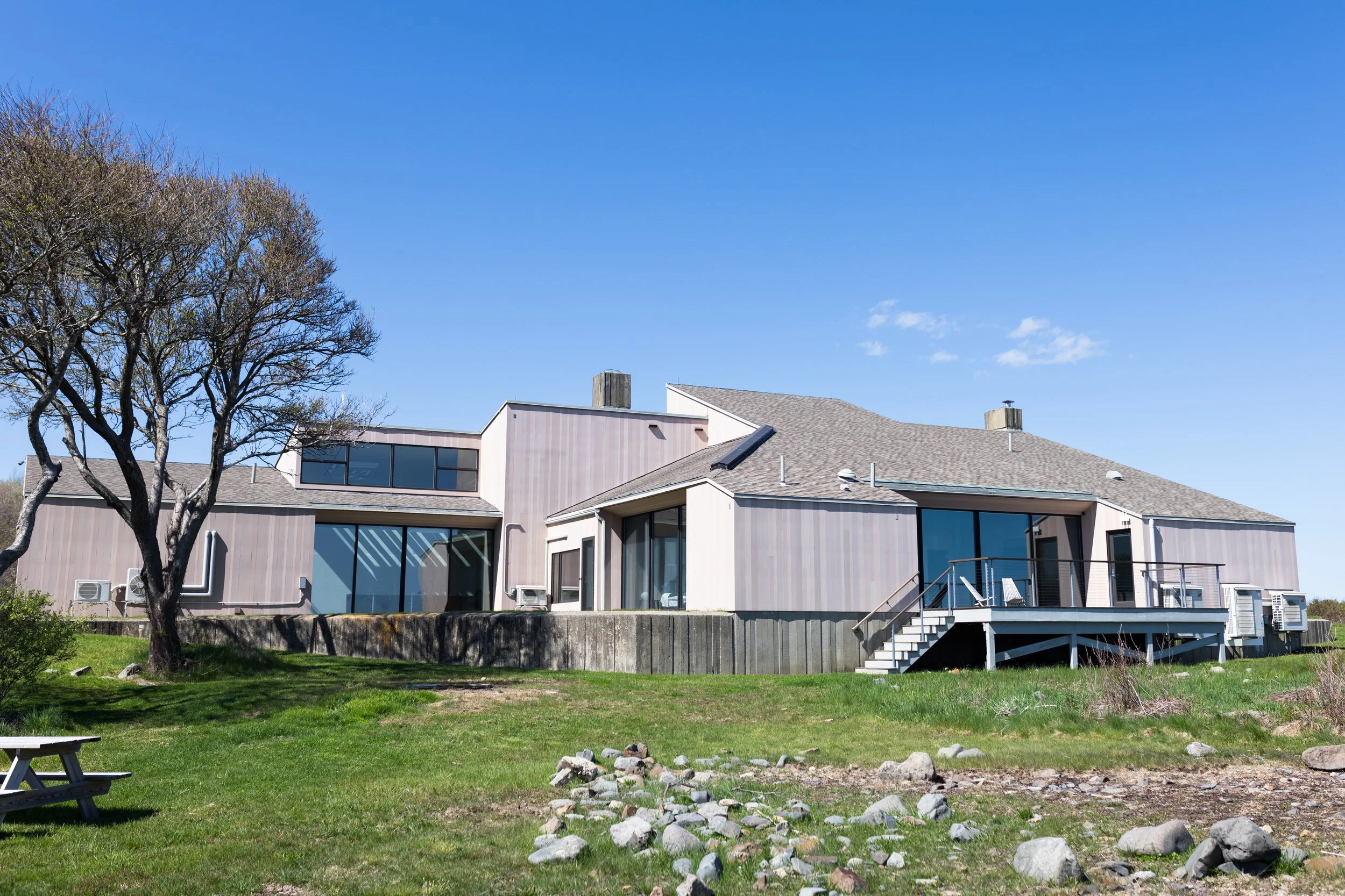 Modern house with large windows and deck, adjacent to a grassy yard with a tree and picnic table under a clear blue sky.
