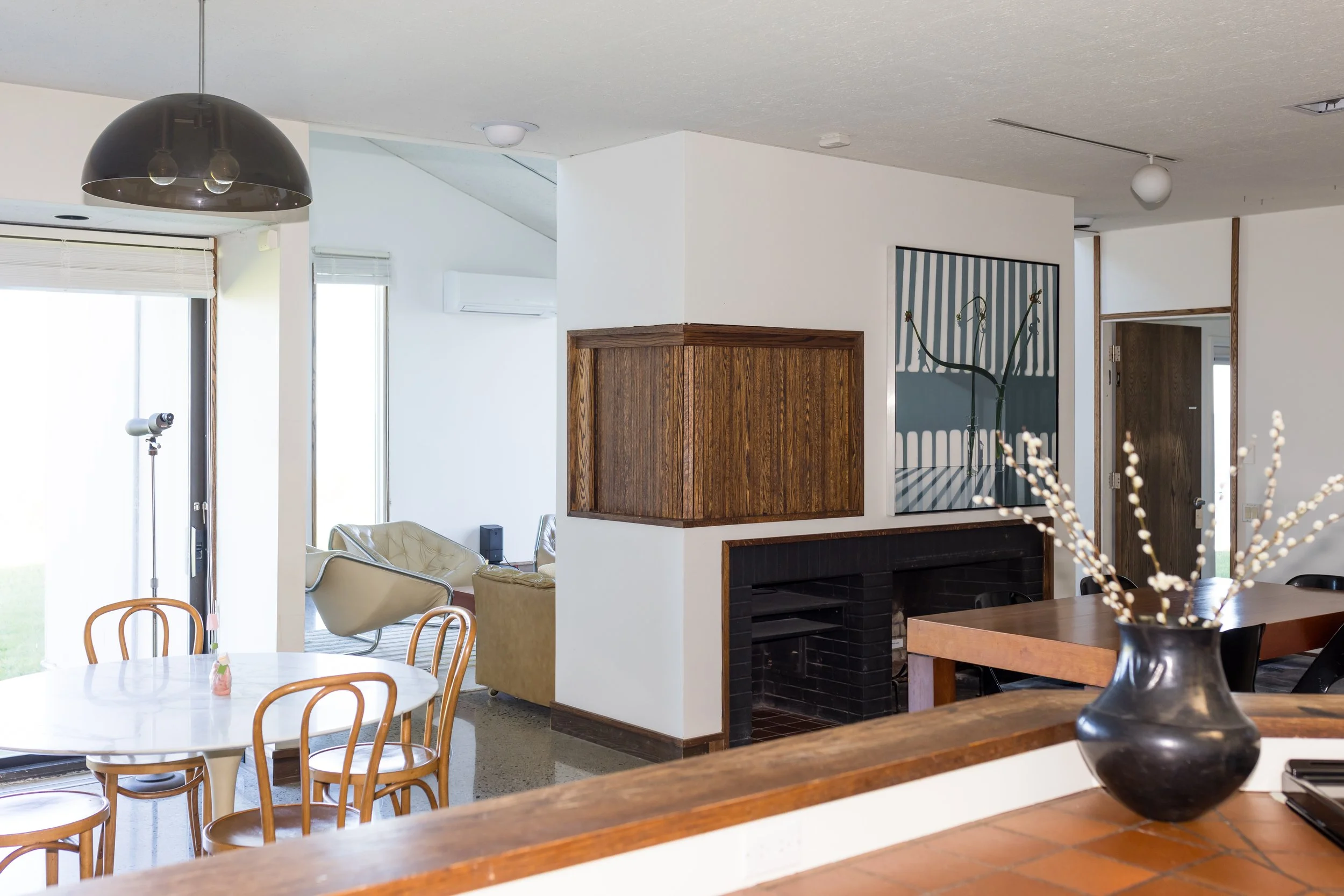 Modern living room with open floor plan, featuring a white marble round table with wooden chairs, black pendant light, black vase with decor, and neutral-toned furniture. A large window and artwork are visible in the background.