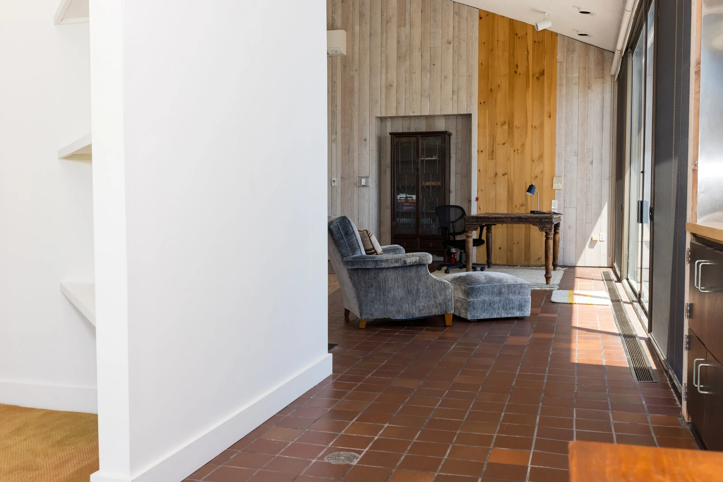 Interior view of a room with brick flooring, a gray armchair, and ottoman. A wooden desk is positioned against a wall with wood paneling. Large windows on the right side allow natural light into the space.
