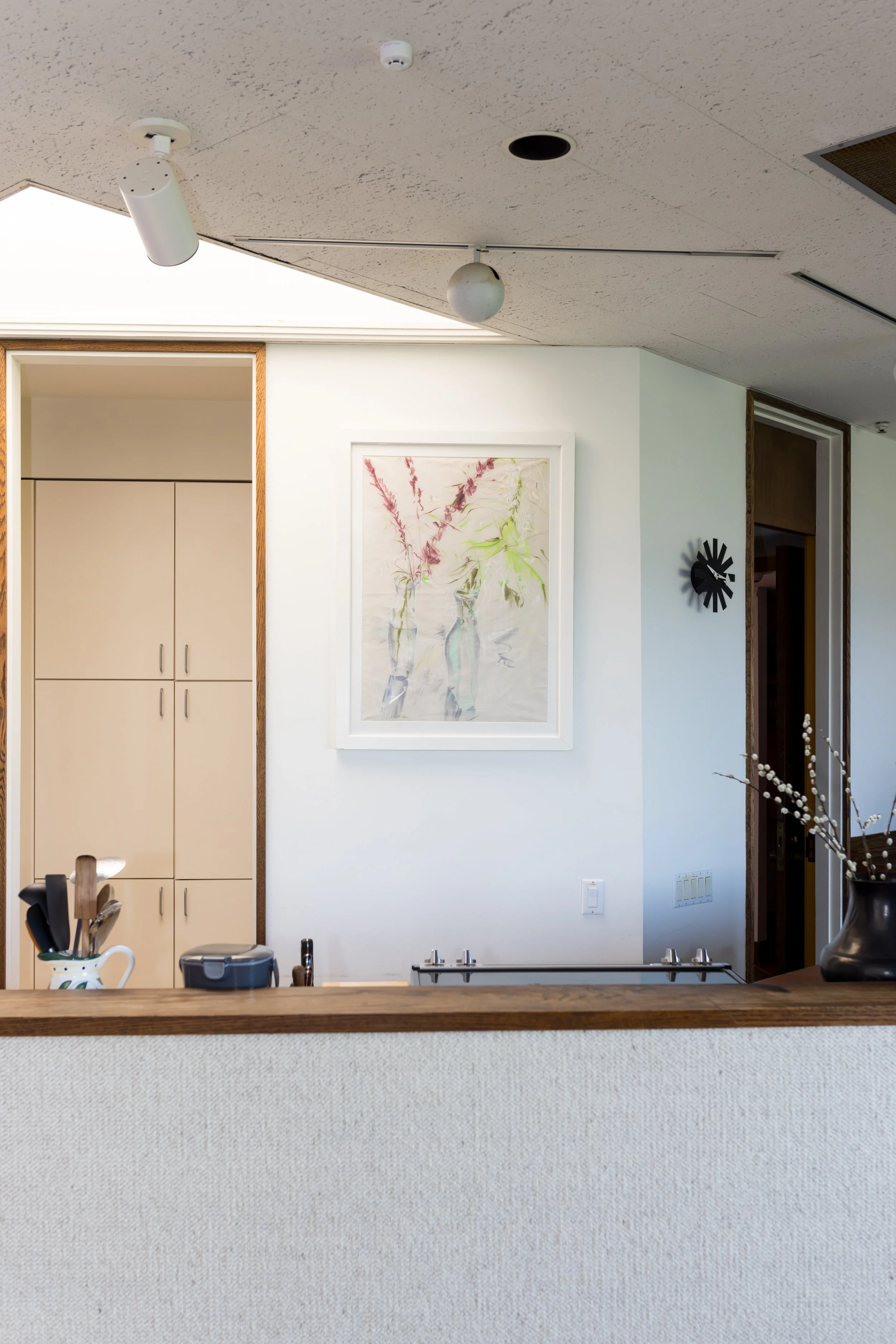 Modern kitchen interior with white cabinetry, wooden countertops, a framed abstract artwork on the wall, utensils in a holder, and decorative branches in a vase.