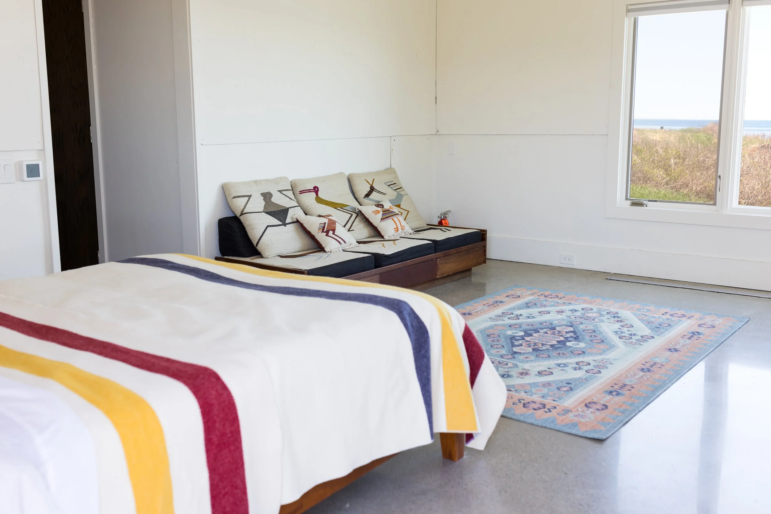 Minimalist bedroom with a bed covered in a striped blanket, a low wooden sofa with artistic cushions, a colorful area rug, and a window with a view of nature.