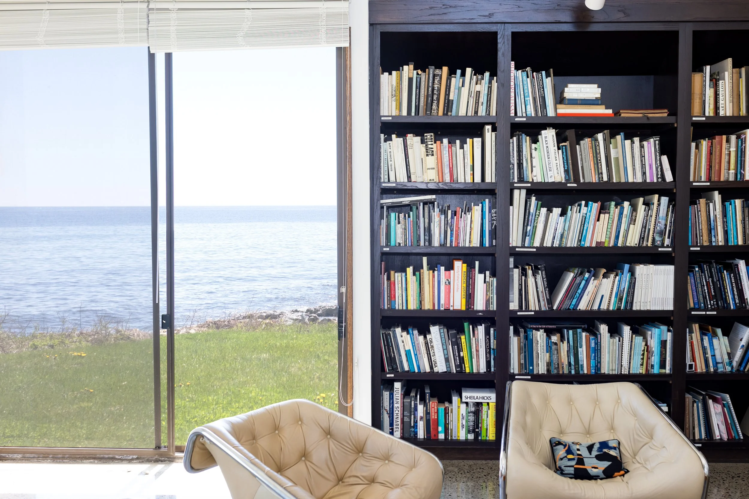 Modern library room with a wall of bookshelves filled with books, a large window with a view of the ocean and grass, and beige armchairs with a colorful pillow.