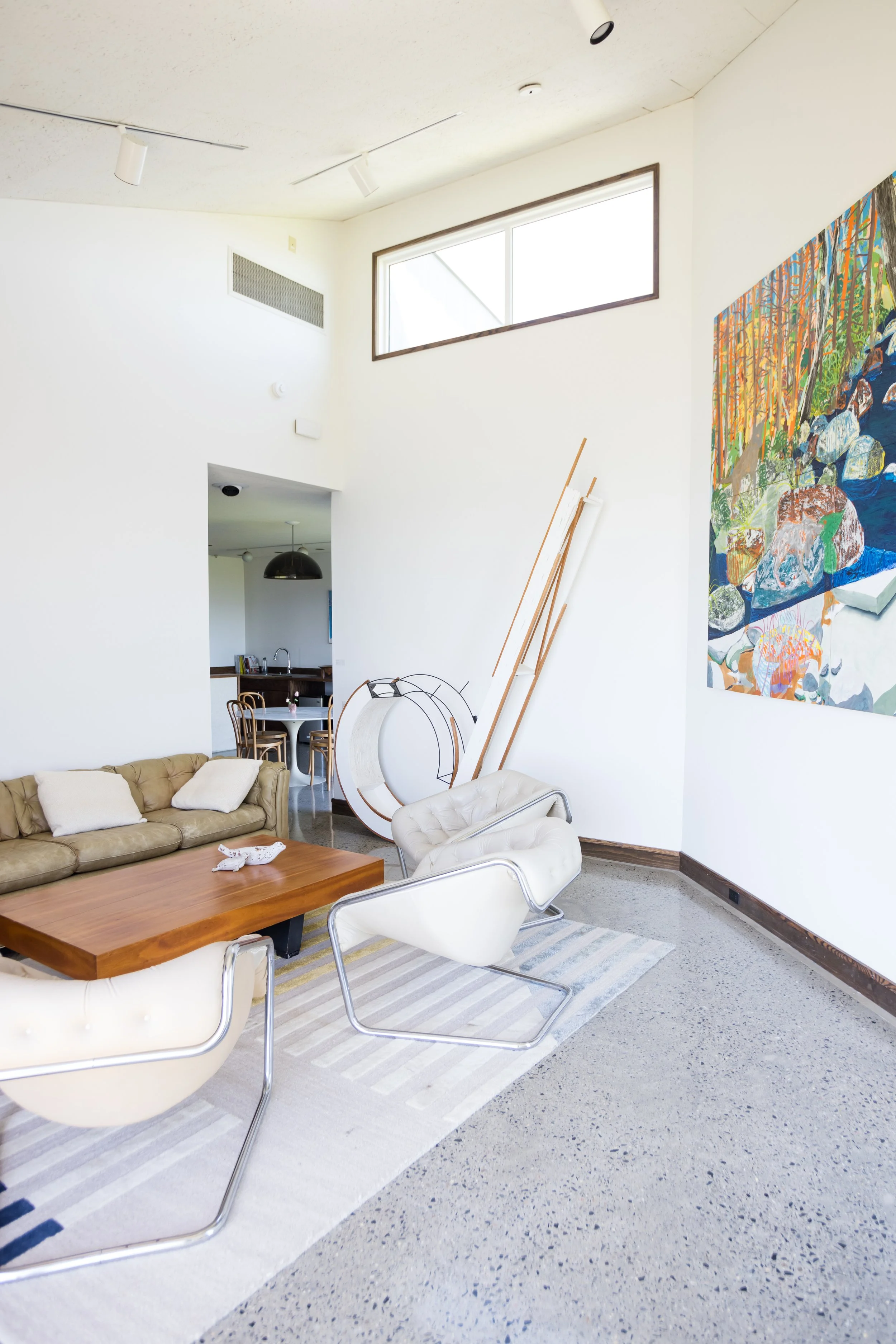Modern living room with beige couch and chairs, wooden coffee table, abstract wall painting, and floor-to-ceiling windows.