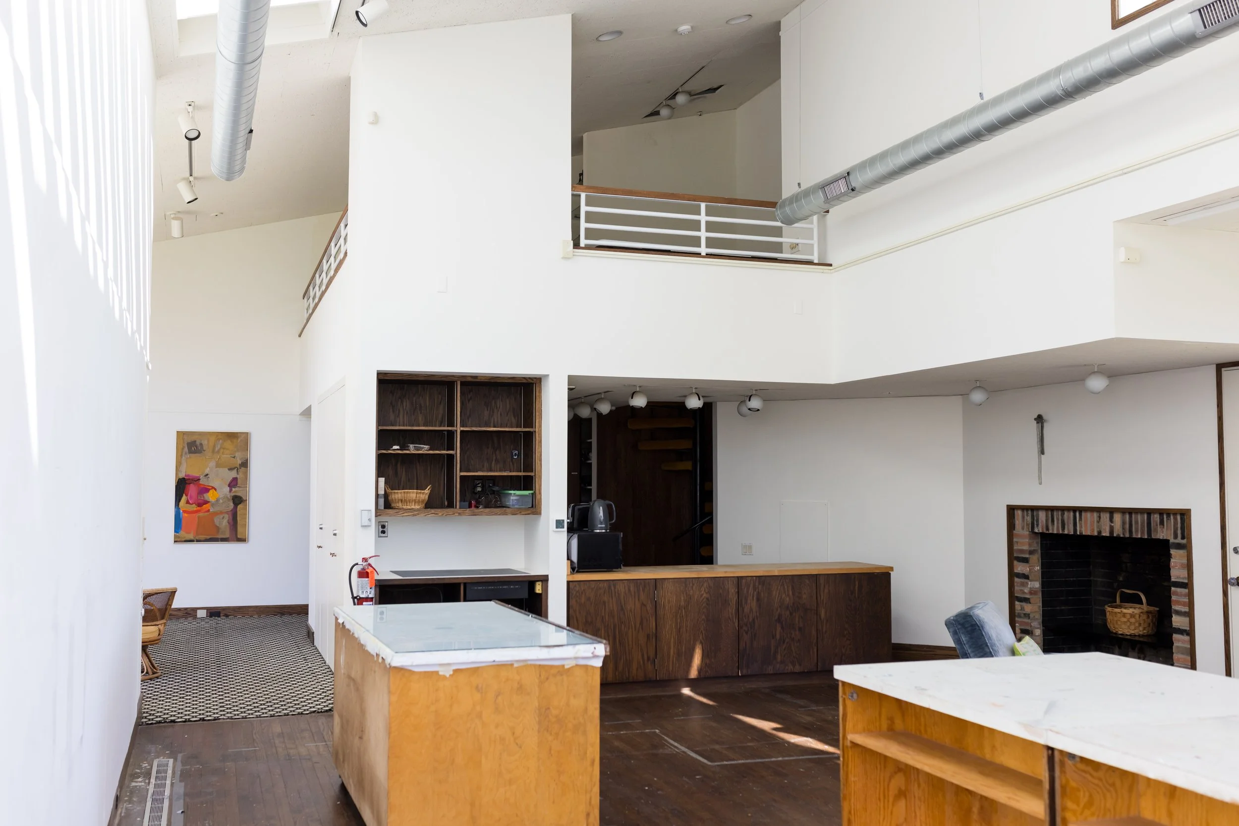Interior of a modern living space featuring white walls, an open kitchen area with wooden cabinets, exposed ductwork, a loft with railing, a brick fireplace, and a piece of abstract art on the wall.