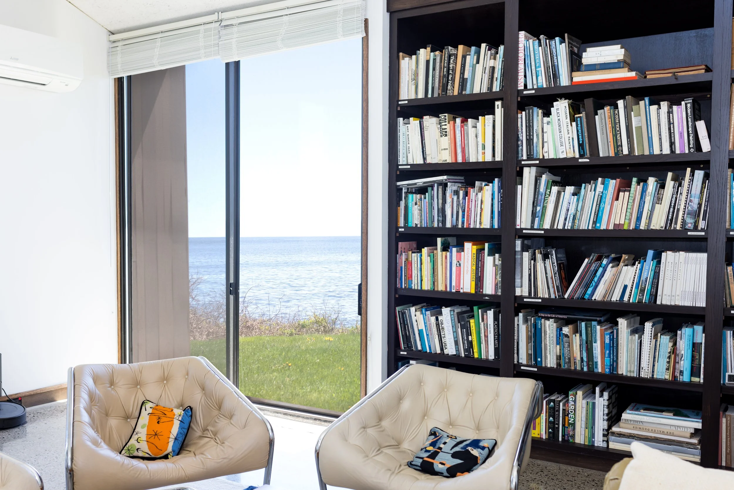 Modern living room with bookshelves, armchairs, and a sea view through large windows.