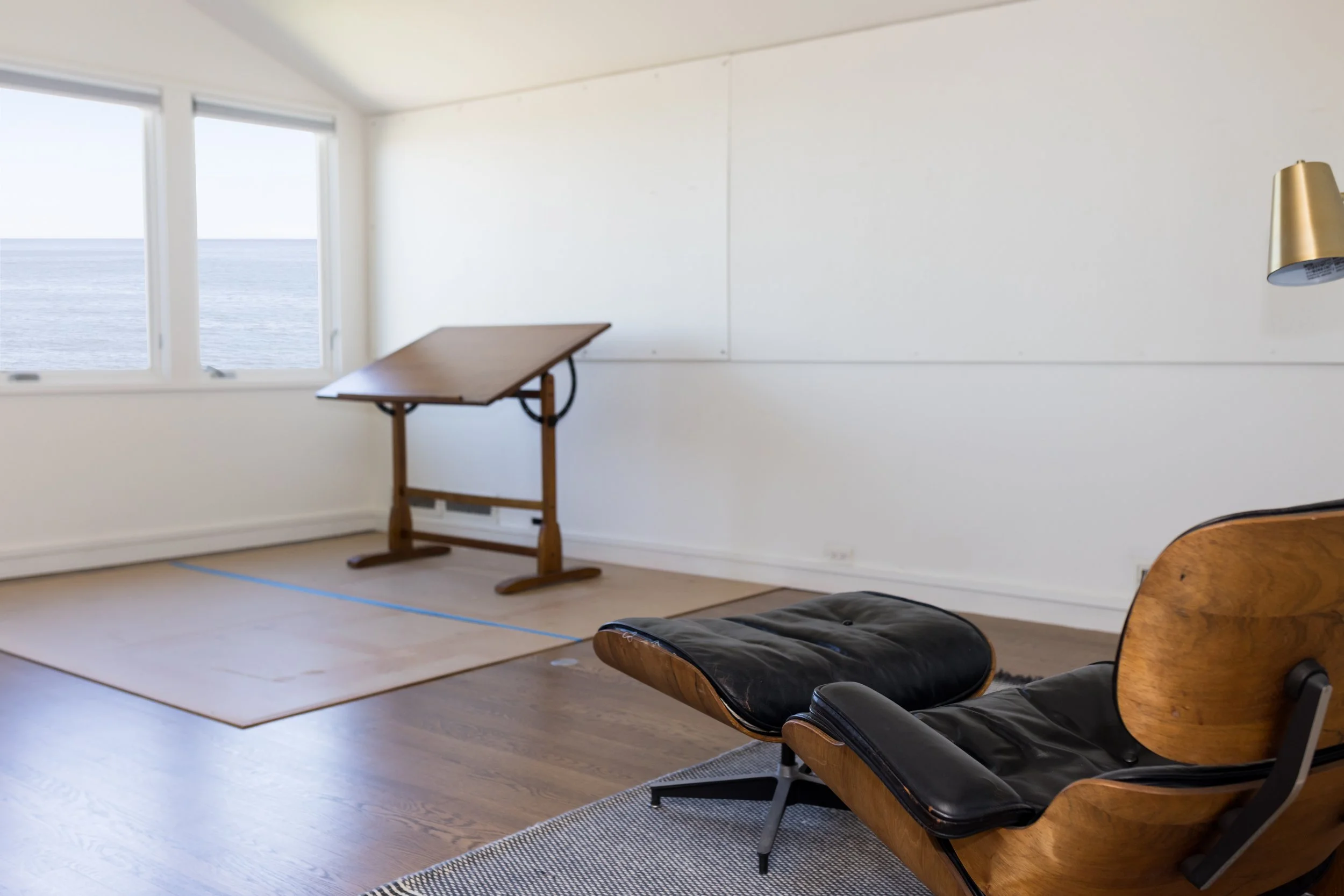Minimalist room with lounge chair, drafting table, large windows overlooking ocean, and wooden floors.