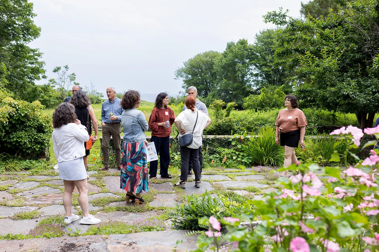 Group of people standing and talking in a garden with green foliage and pink flowers.